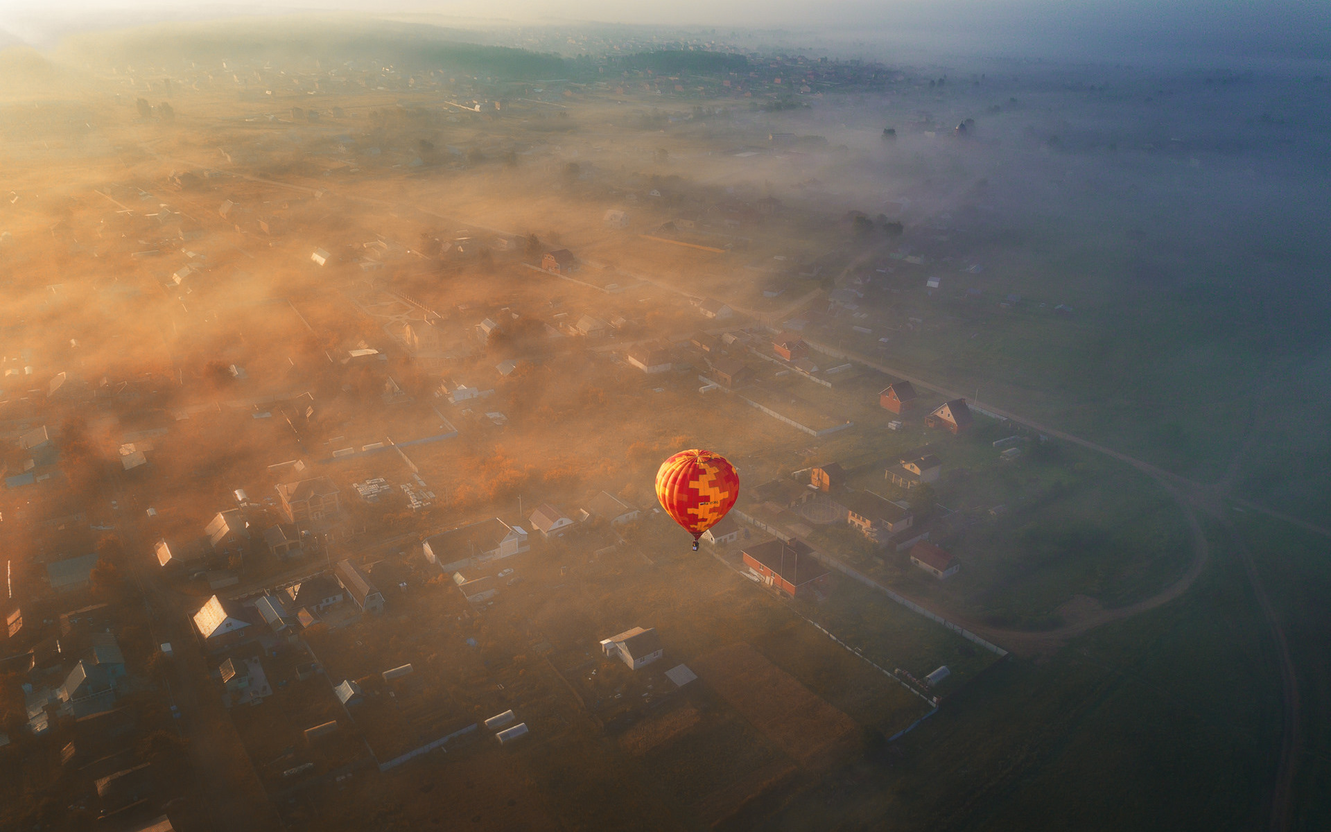 Aerial view of a hot air balloon over a misty village at sunrise HD Wallpaper