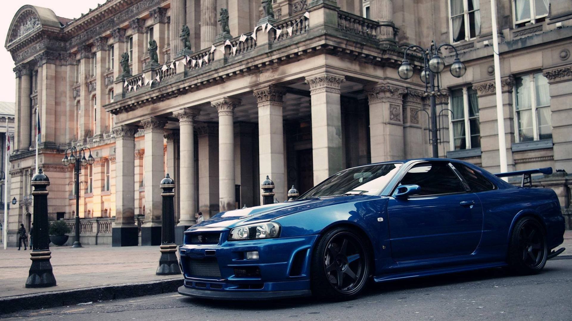 Blue Nissan Skyline R34 GT-R in front of a building HD Wallpaper