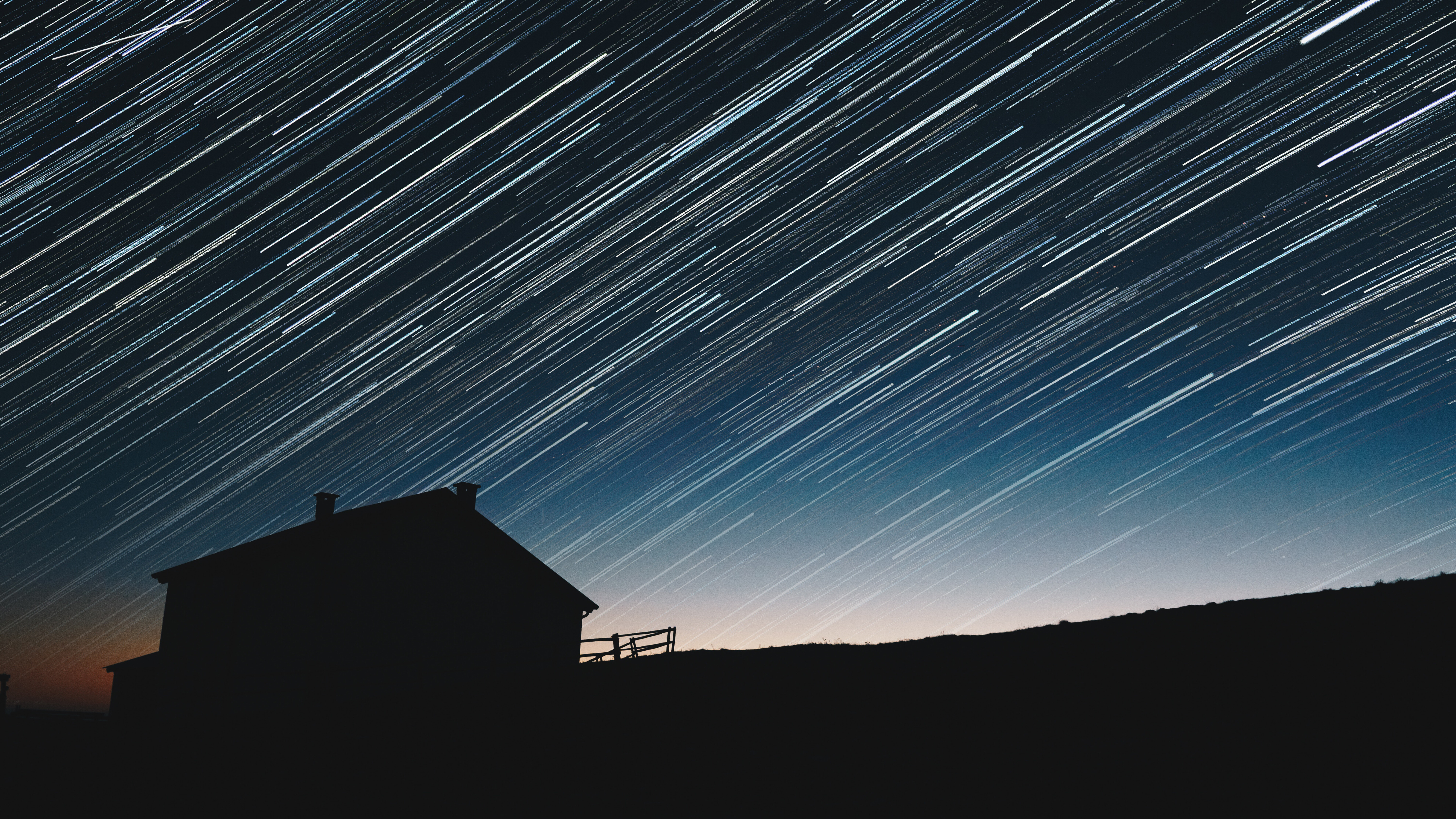 Star trails over a silhouetted house and hill HD Wallpaper