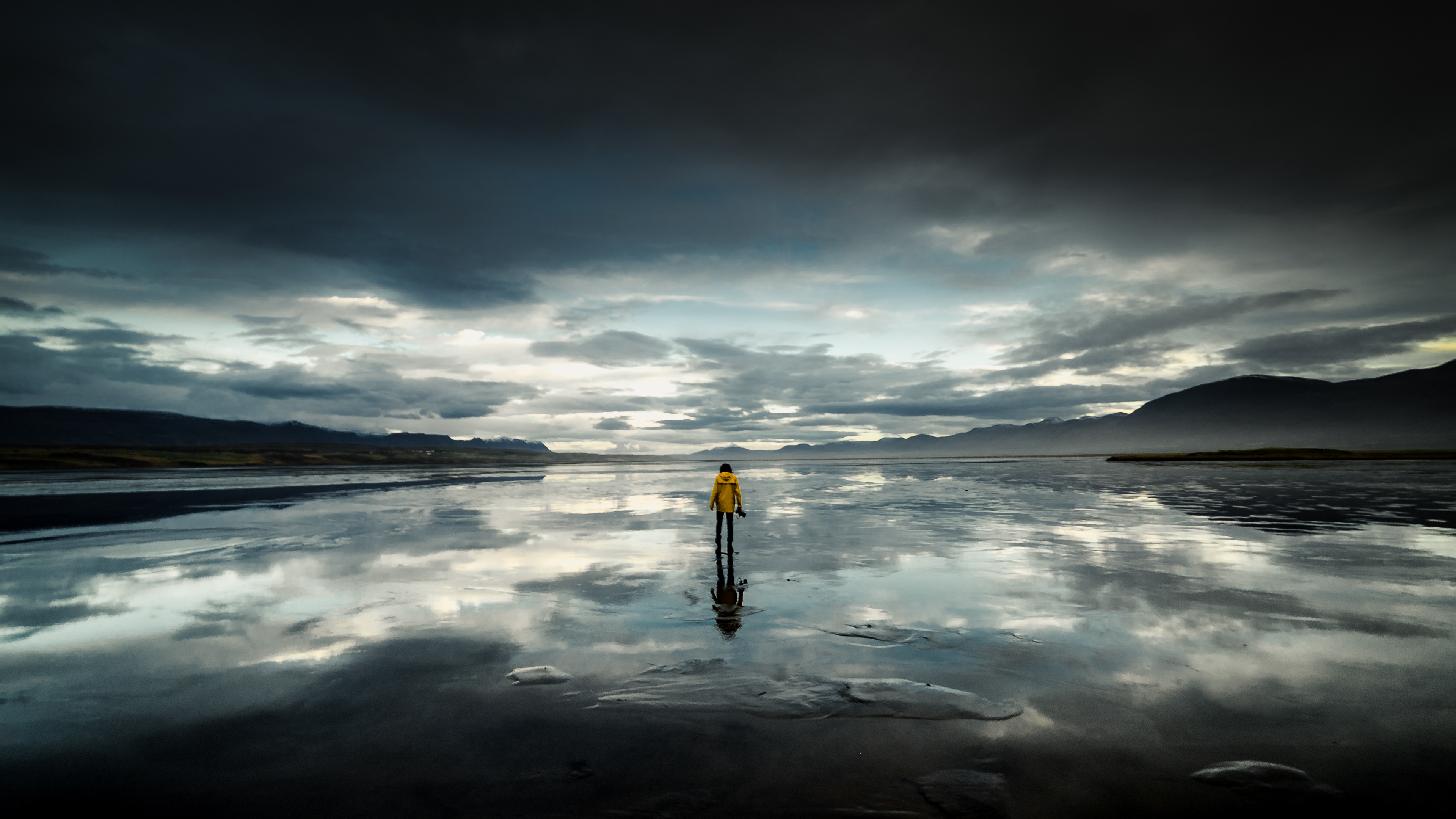 Person in yellow jacket on reflective beach under cloudy sky 4K HD Wallpaper