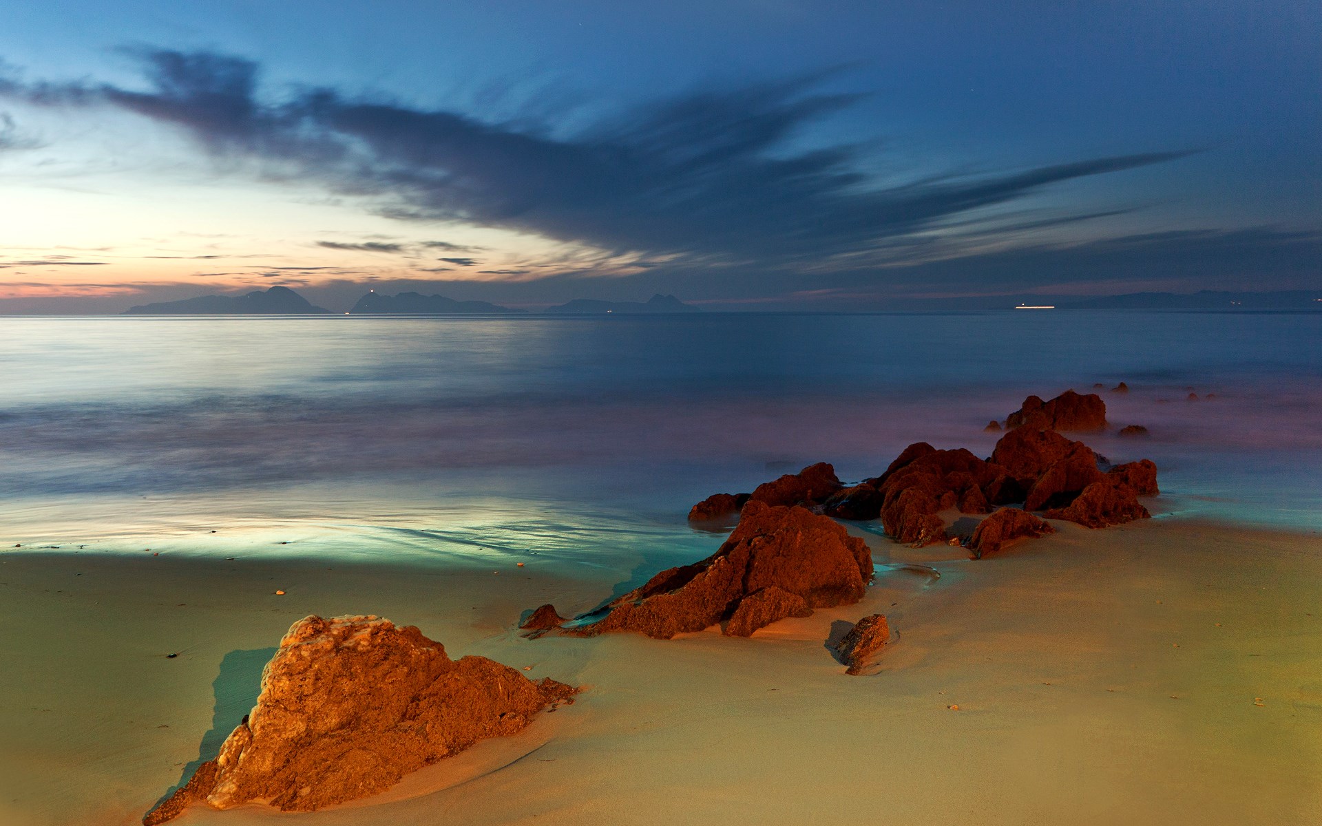 Beach at dusk with rocks and distant islands HD Wallpaper
