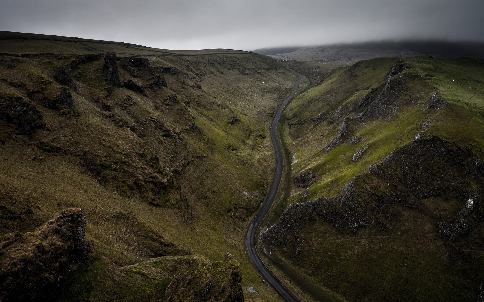 Winding road through a green valley under a cloudy sky HD Wallpaper