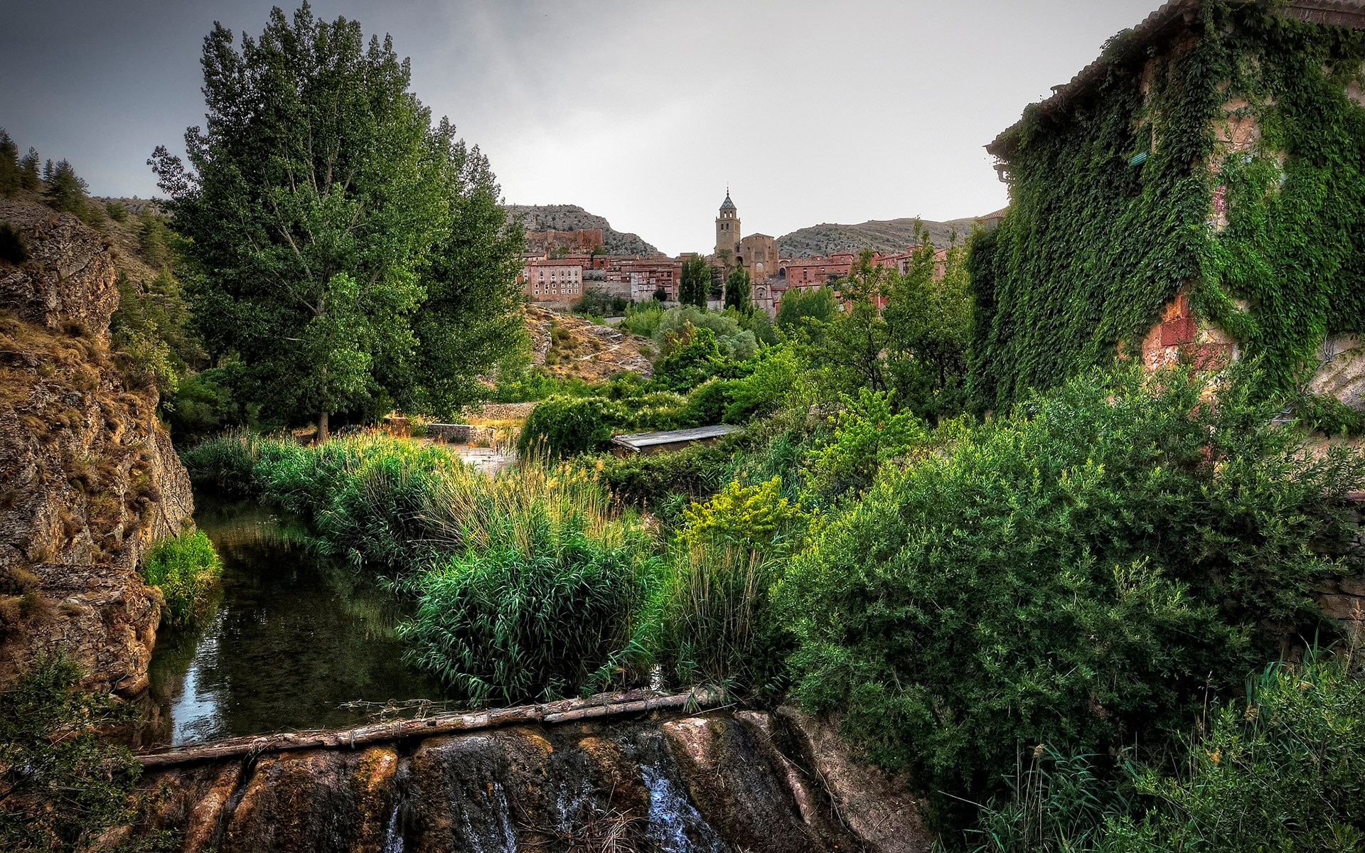 Village with a river and overgrown building HD Wallpaper