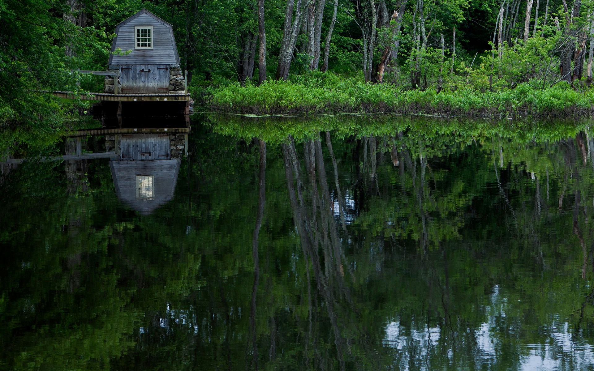 Wooden boathouse reflected in a calm lake surrounded by trees HD Wallpaper