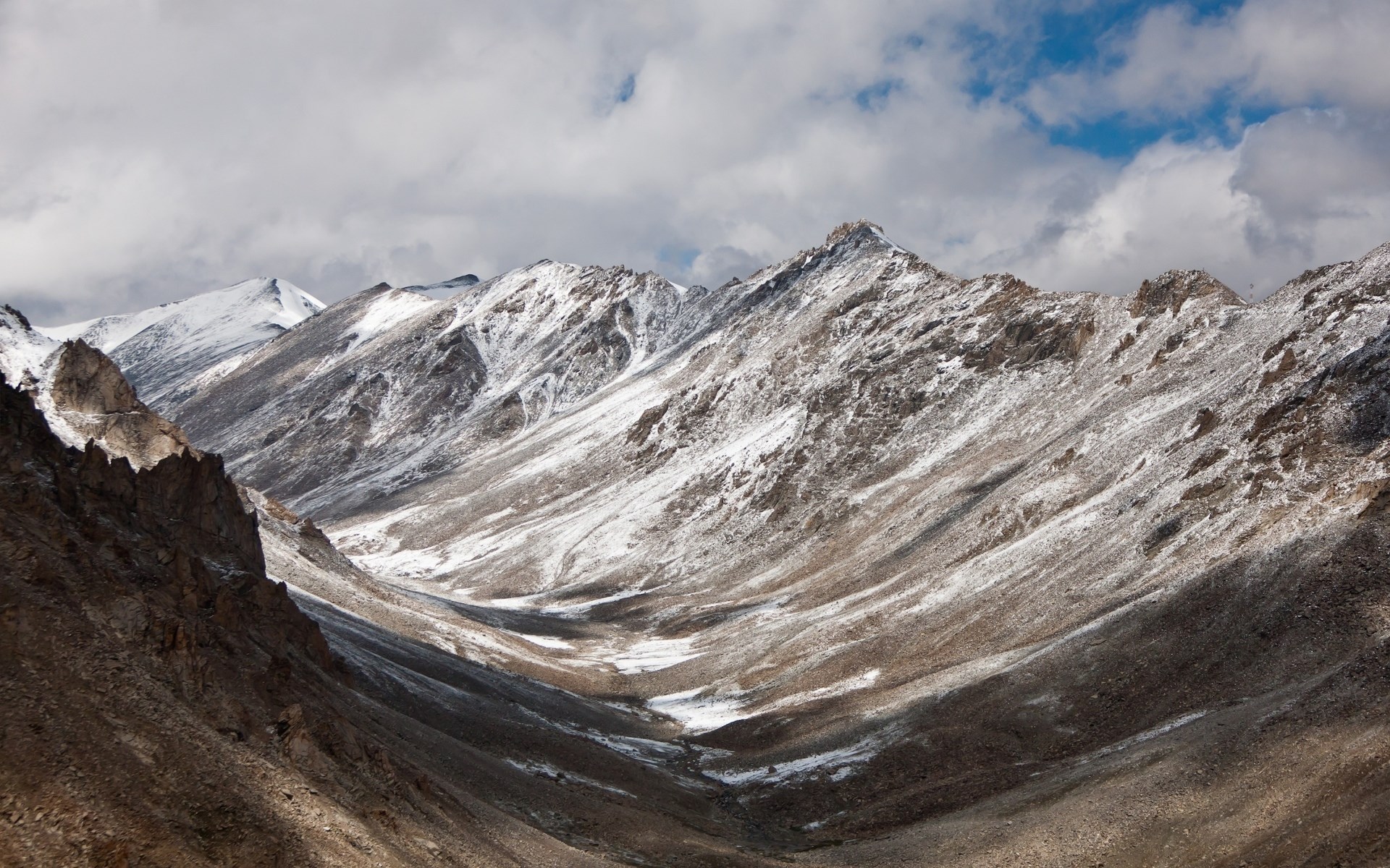Snow-dusted mountains in a valley under a cloudy sky HD Wallpaper