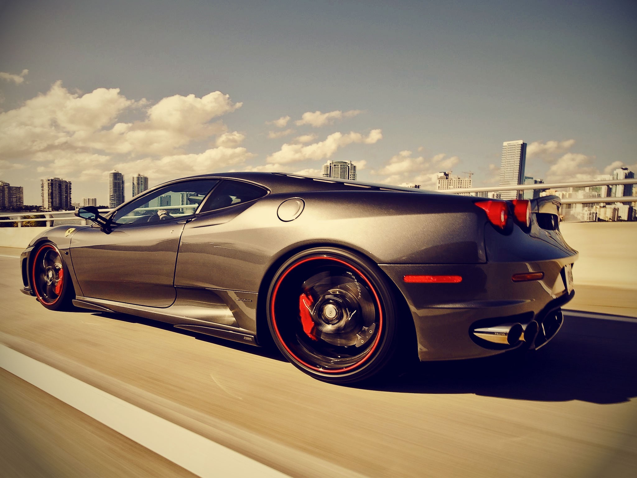 Gray Ferrari F430 driving on a highway with city skyline HD Wallpaper