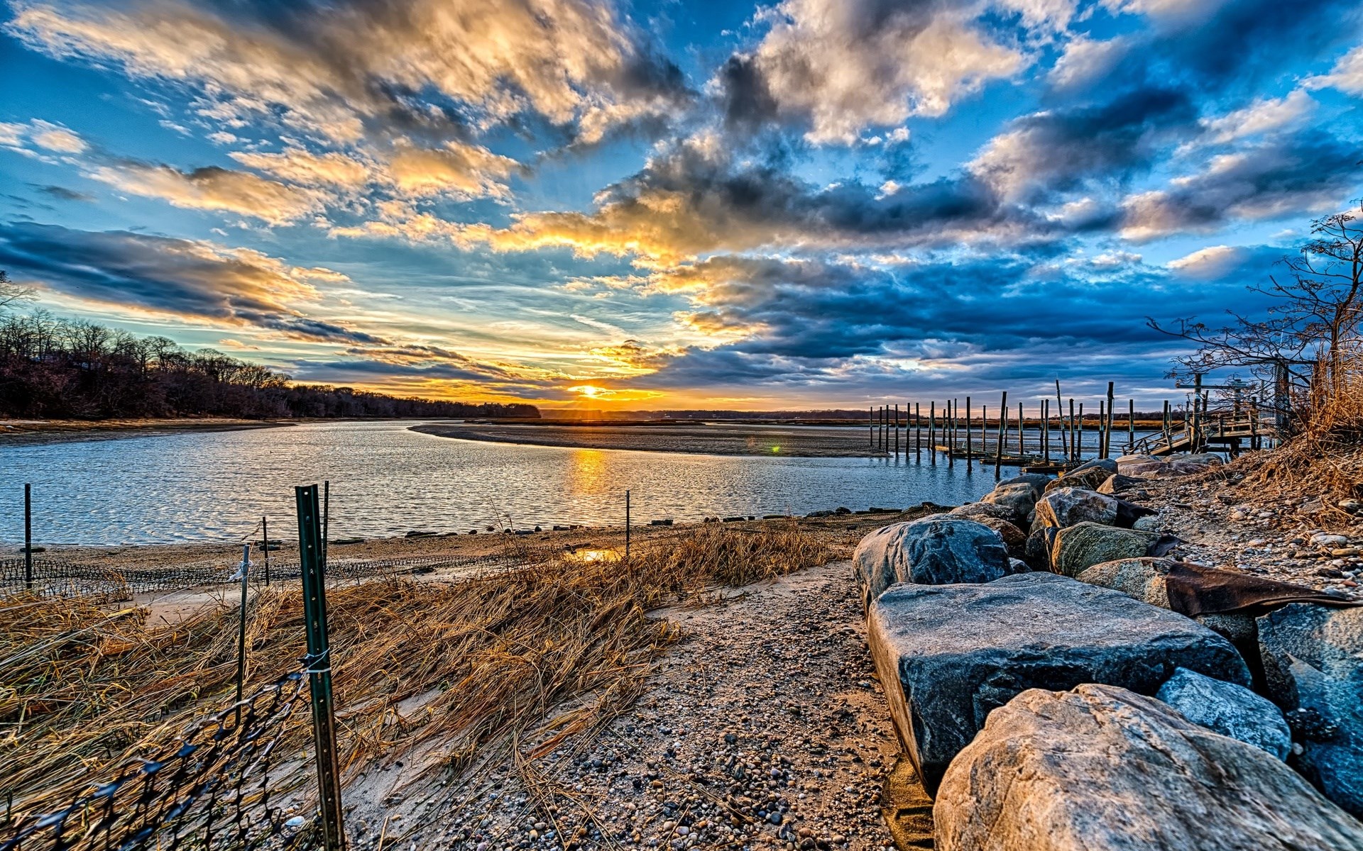 Sunset over a calm river with a wooden pier and rocky shore HD Wallpaper