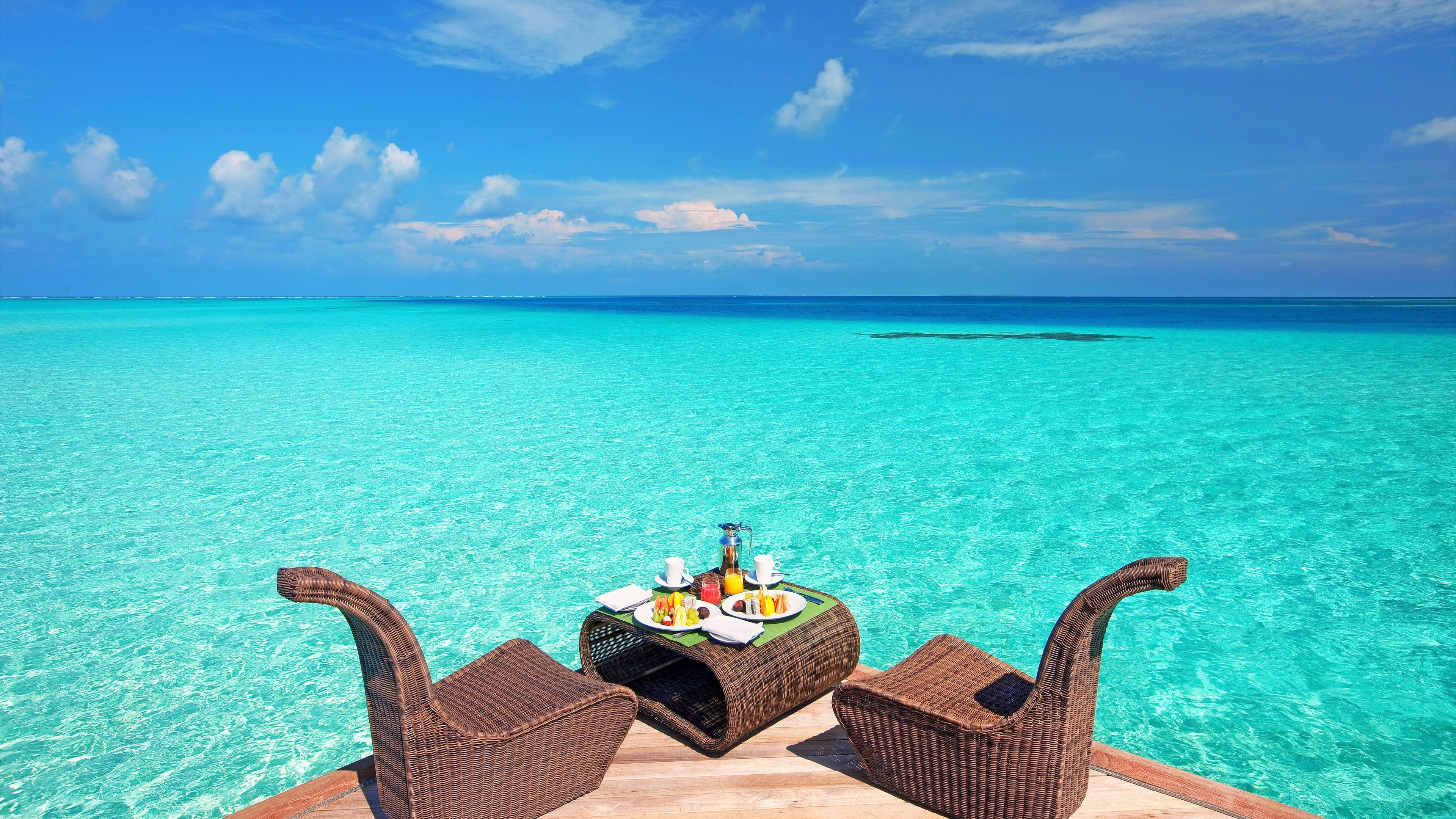 Breakfast table set for two on a wooden deck overlooking a turquoise ocean HD Wallpaper