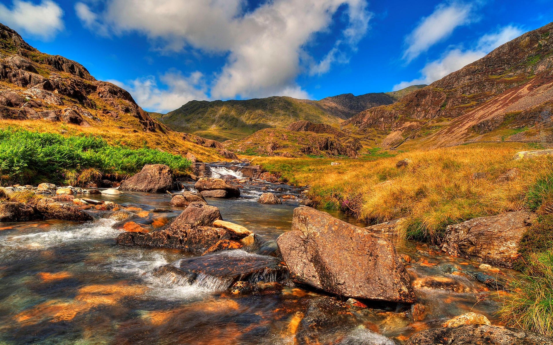 Mountain stream flowing through rocky terrain under a blue sky HD Wallpaper