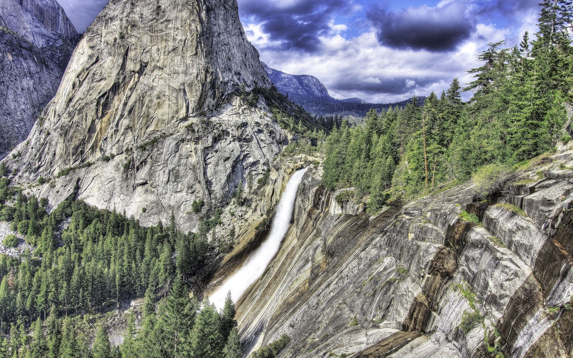 Waterfall cascading down a rocky mountain with pine trees HD Wallpaper
