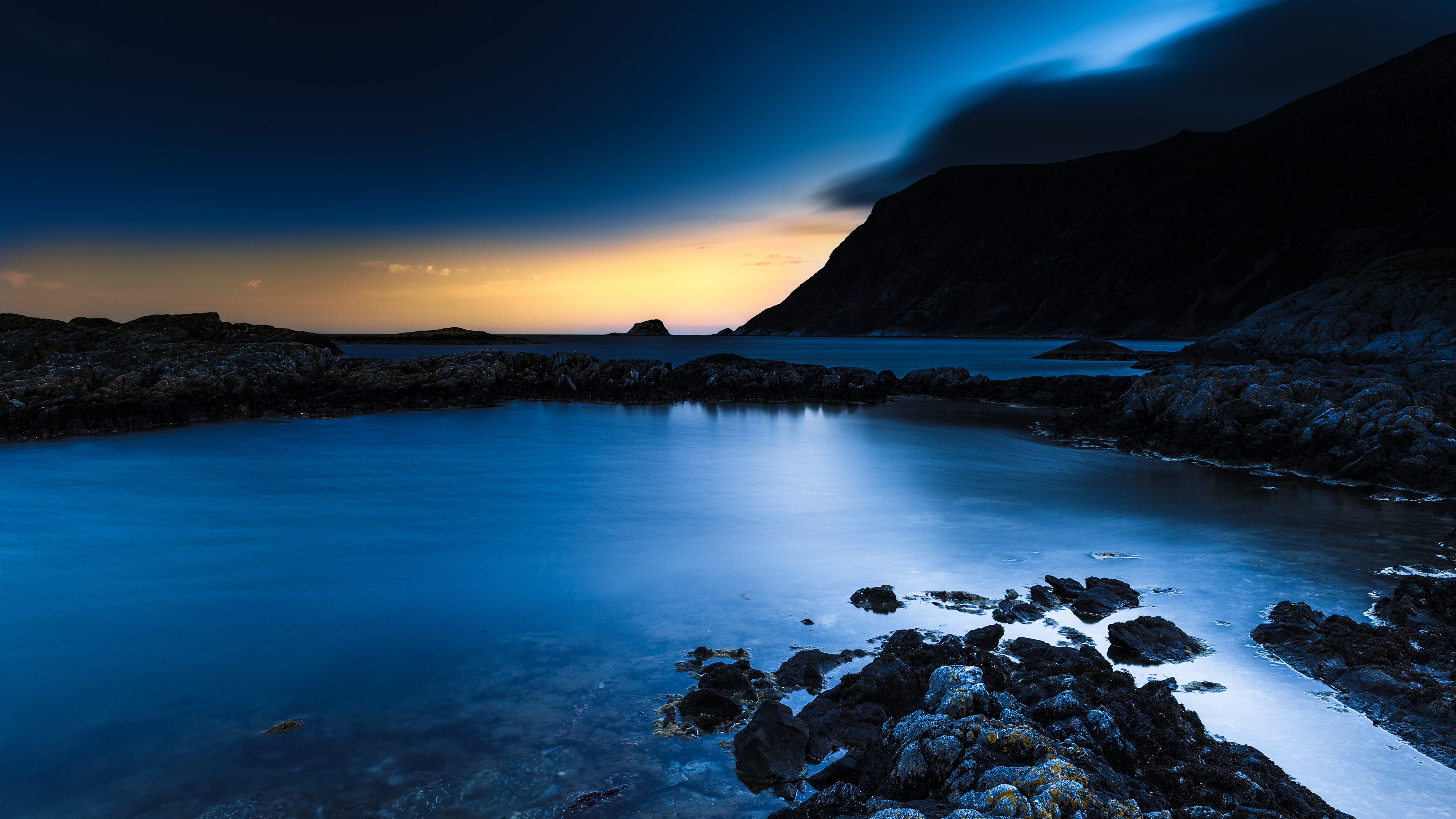 Dark rocky coastline with calm blue water at dusk HD Wallpaper