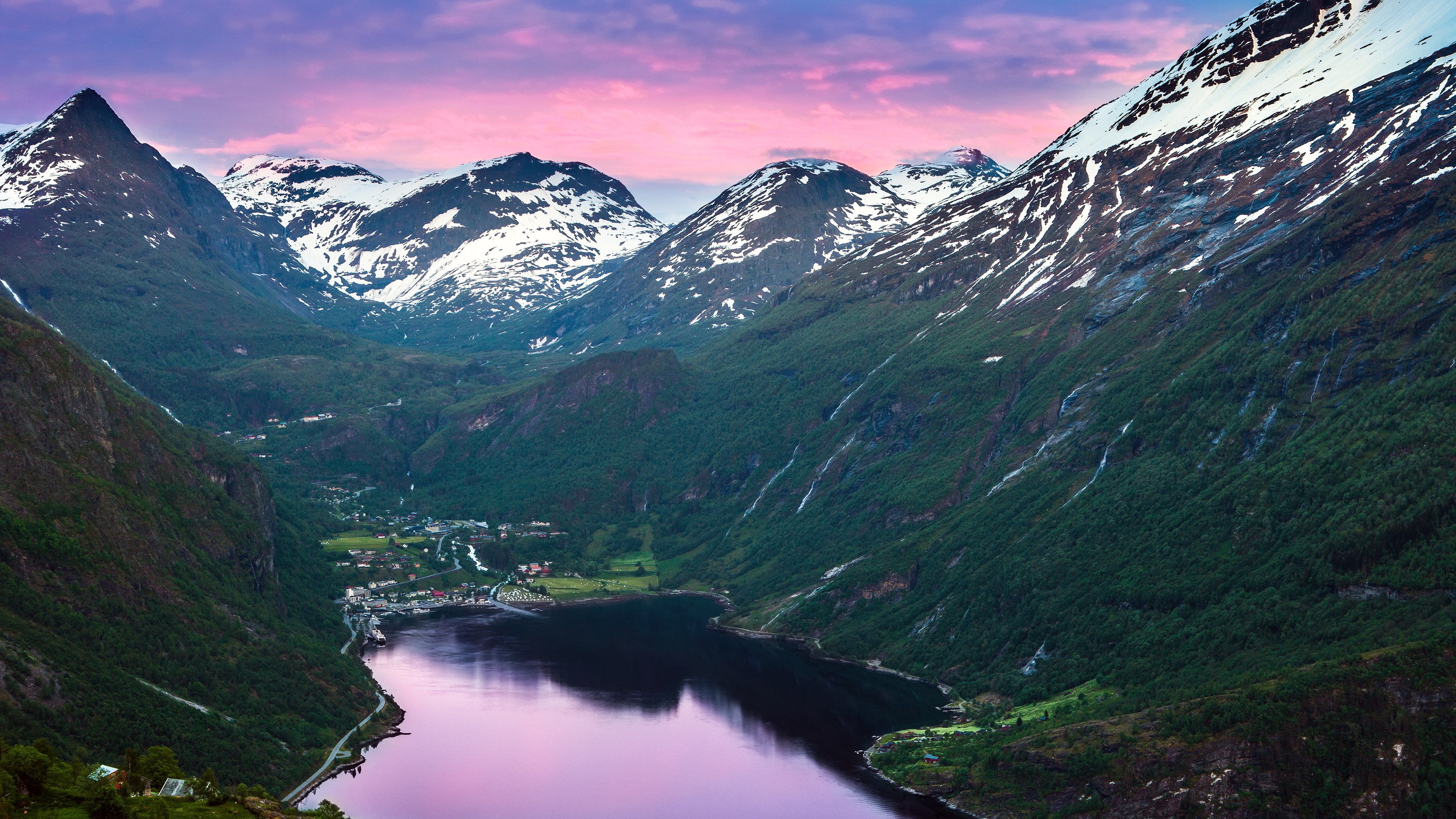 Snow-capped mountains overlook a fjord and village at sunset HD Wallpaper