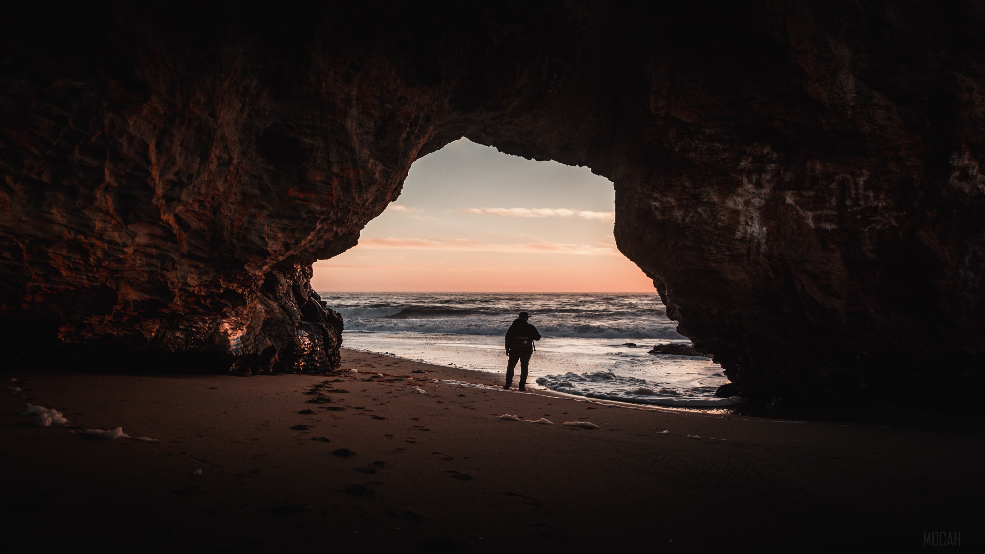 Person standing in cave entrance at sunset HD Wallpaper