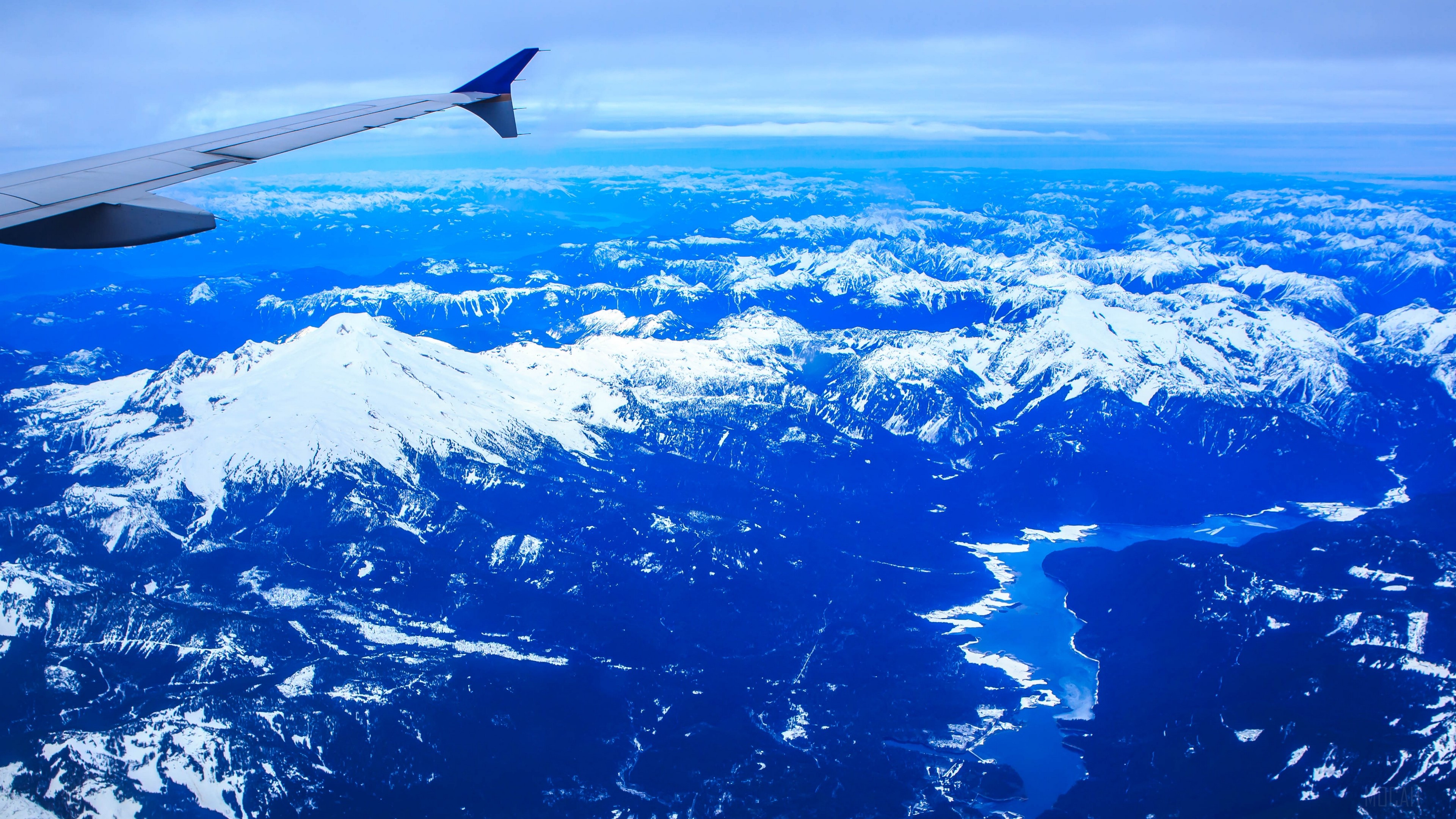 Airplane wing over snow-capped mountains and lake HD Wallpaper