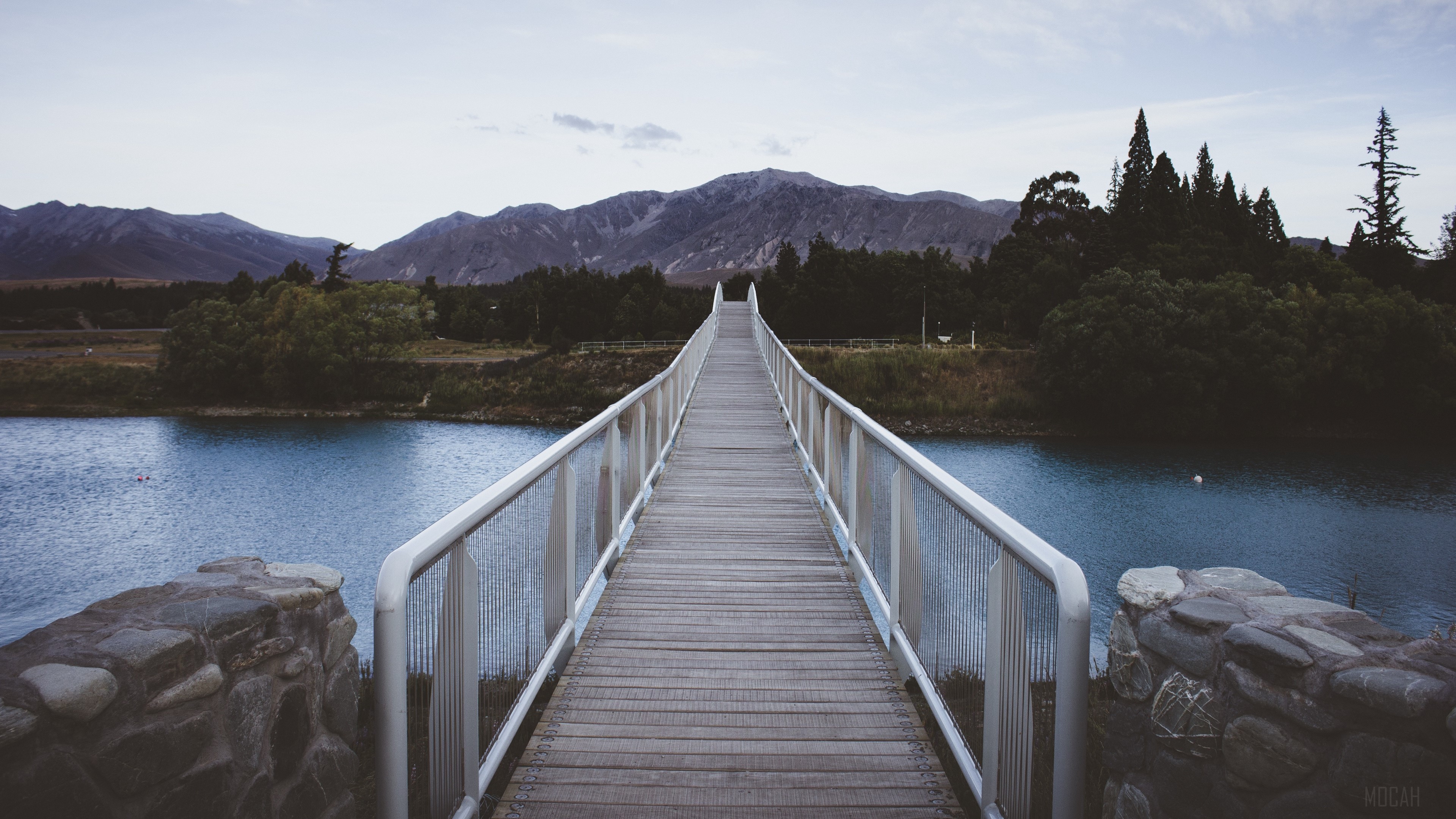 Wooden bridge over a blue river with mountains in the background 4K HD Wallpaper