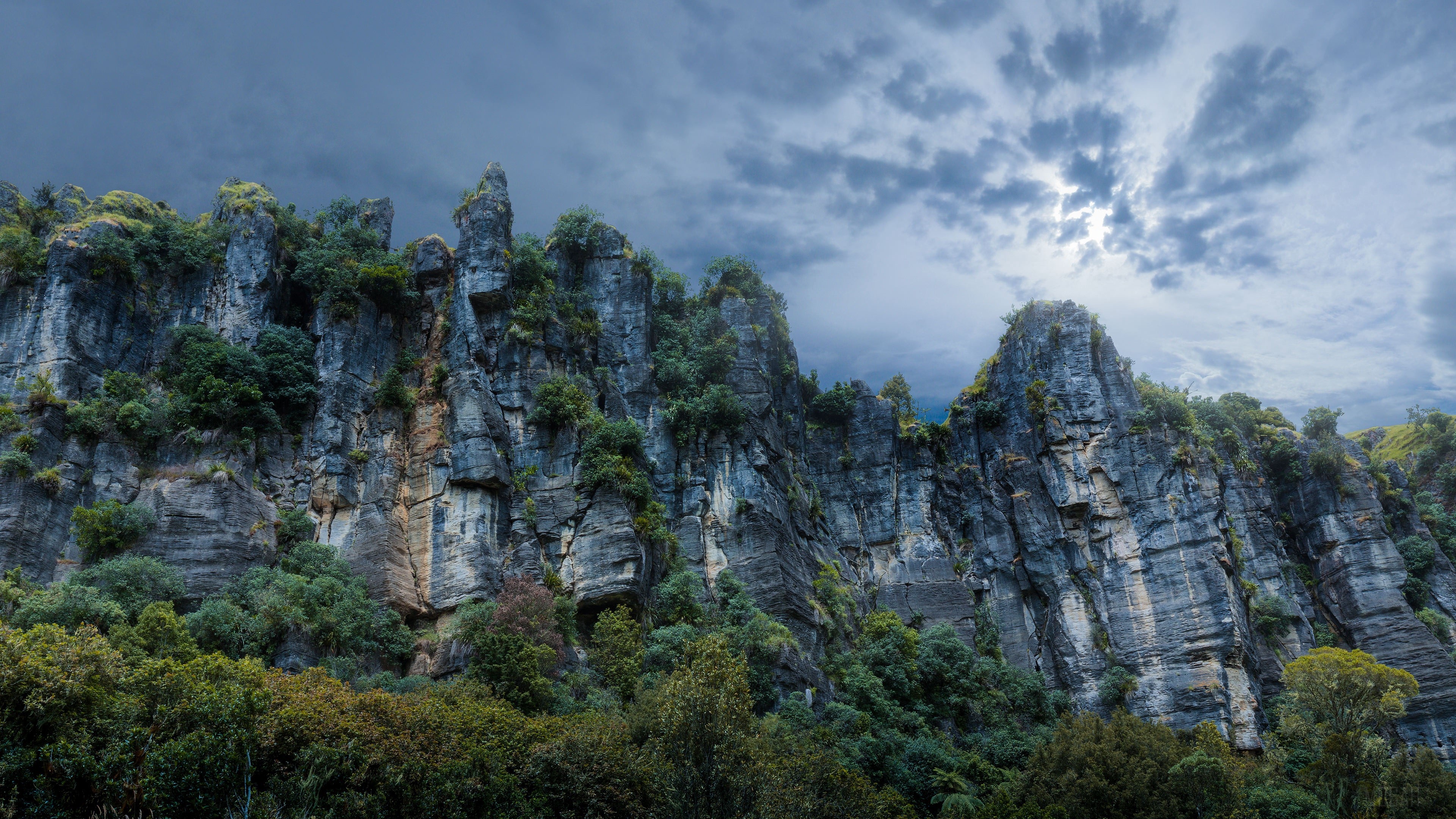 Rocky cliffs with trees under a cloudy sky HD Wallpaper