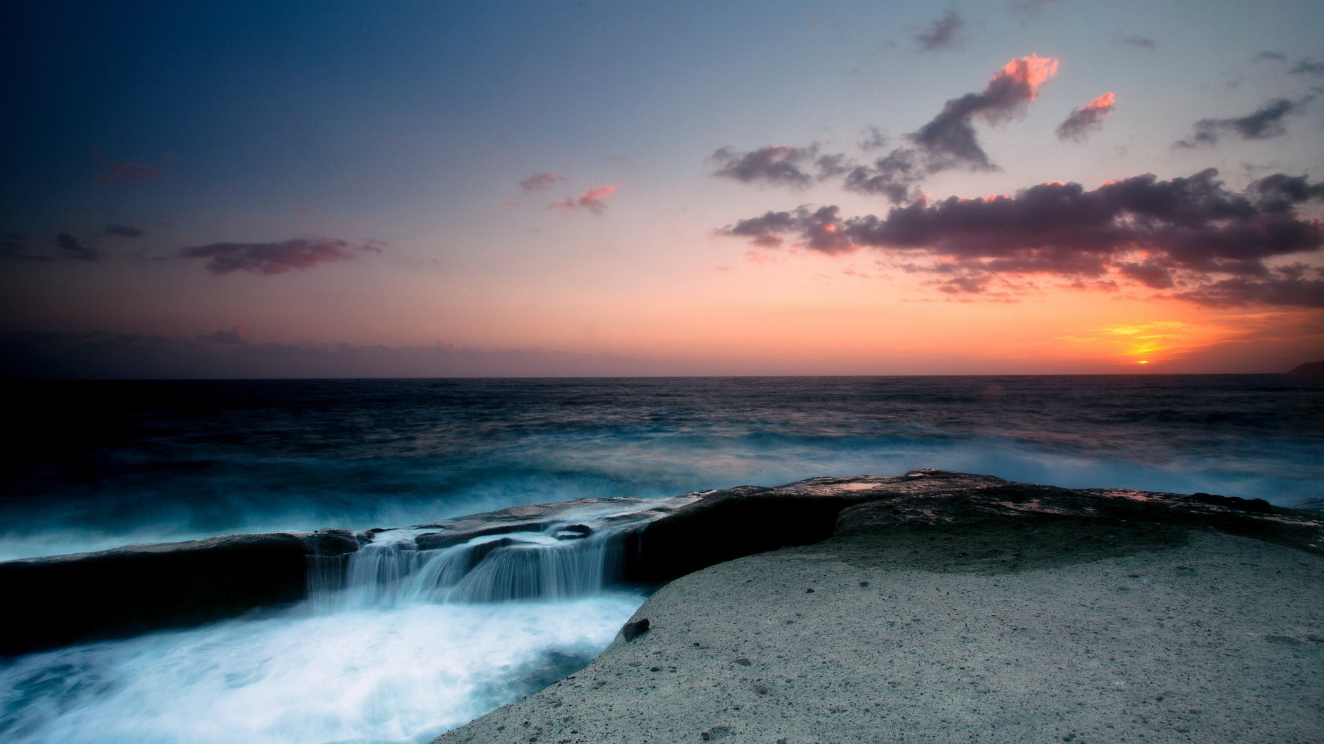Sunset over the ocean with waves crashing on rocks HD Wallpaper