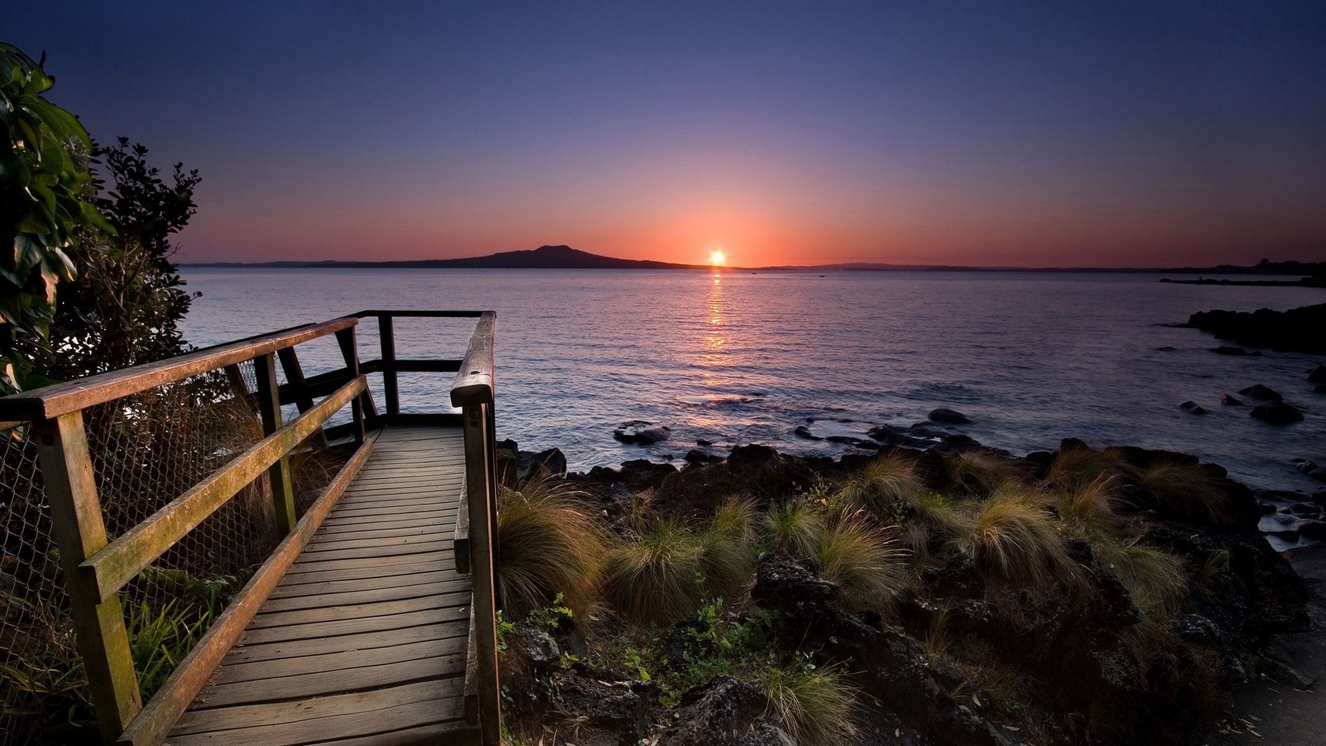 Wooden walkway overlooking ocean at sunset with island HD Wallpaper