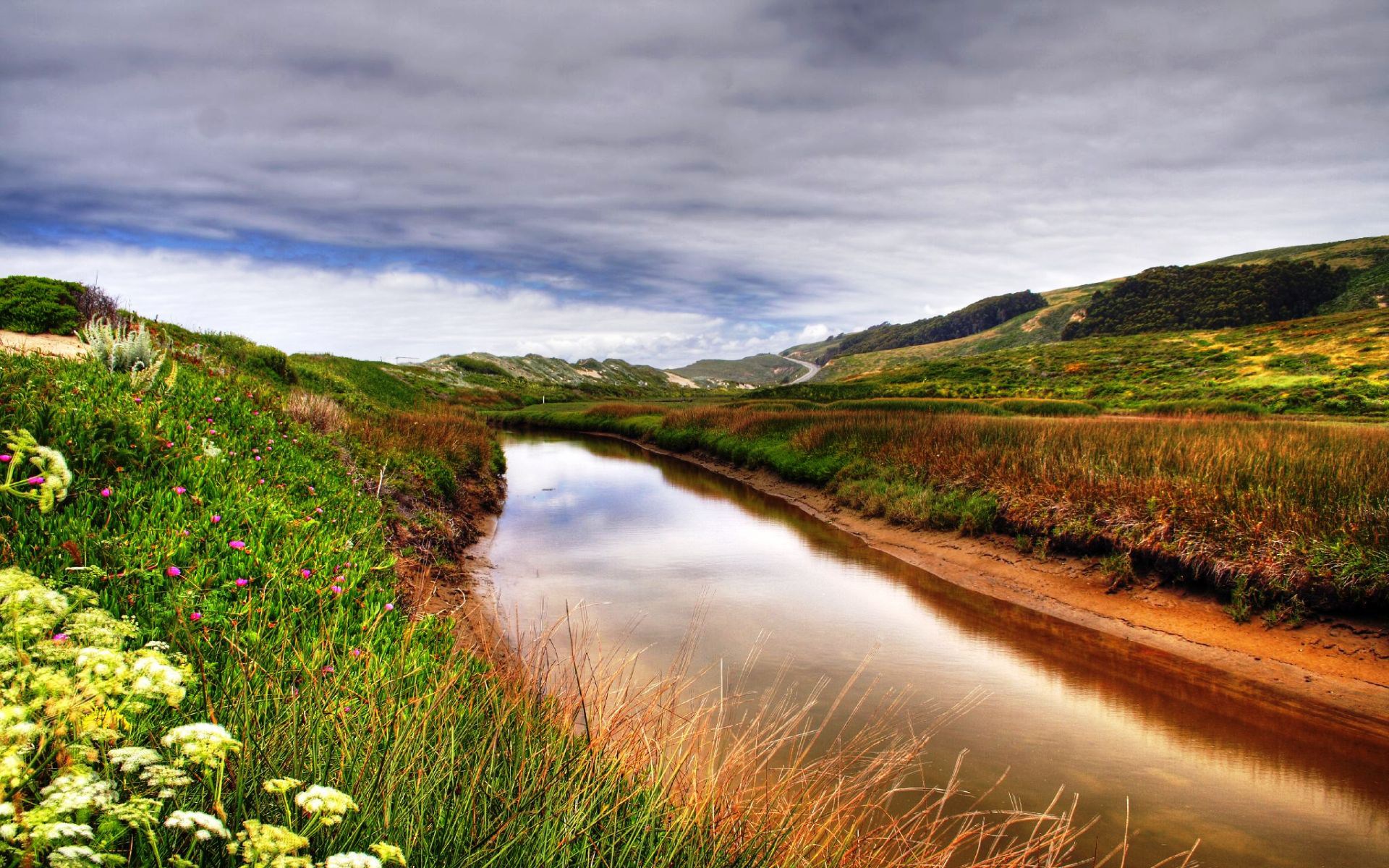 A winding river flows through a grassy landscape under a cloudy sky. HD Wallpaper