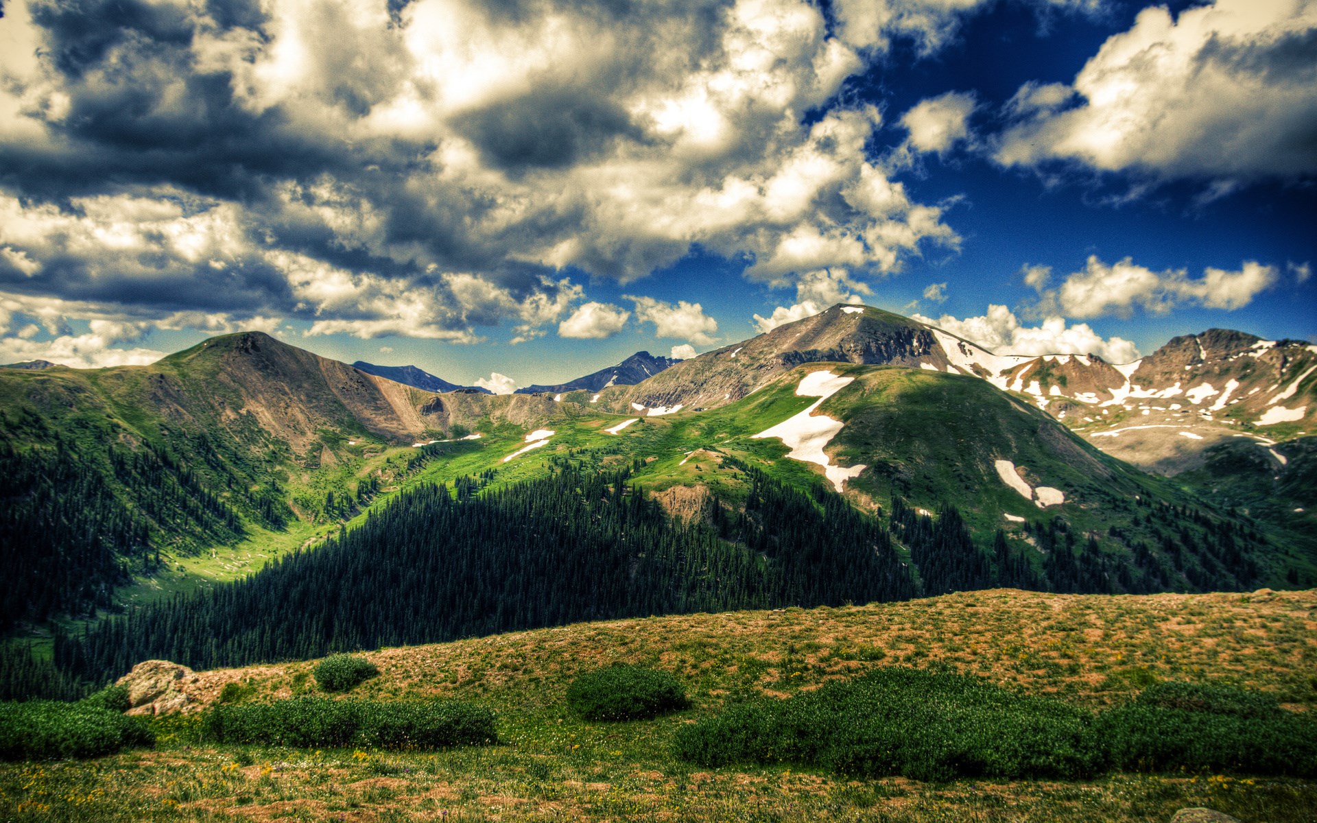 Green mountains with snow patches under a cloudy sky HD Wallpaper