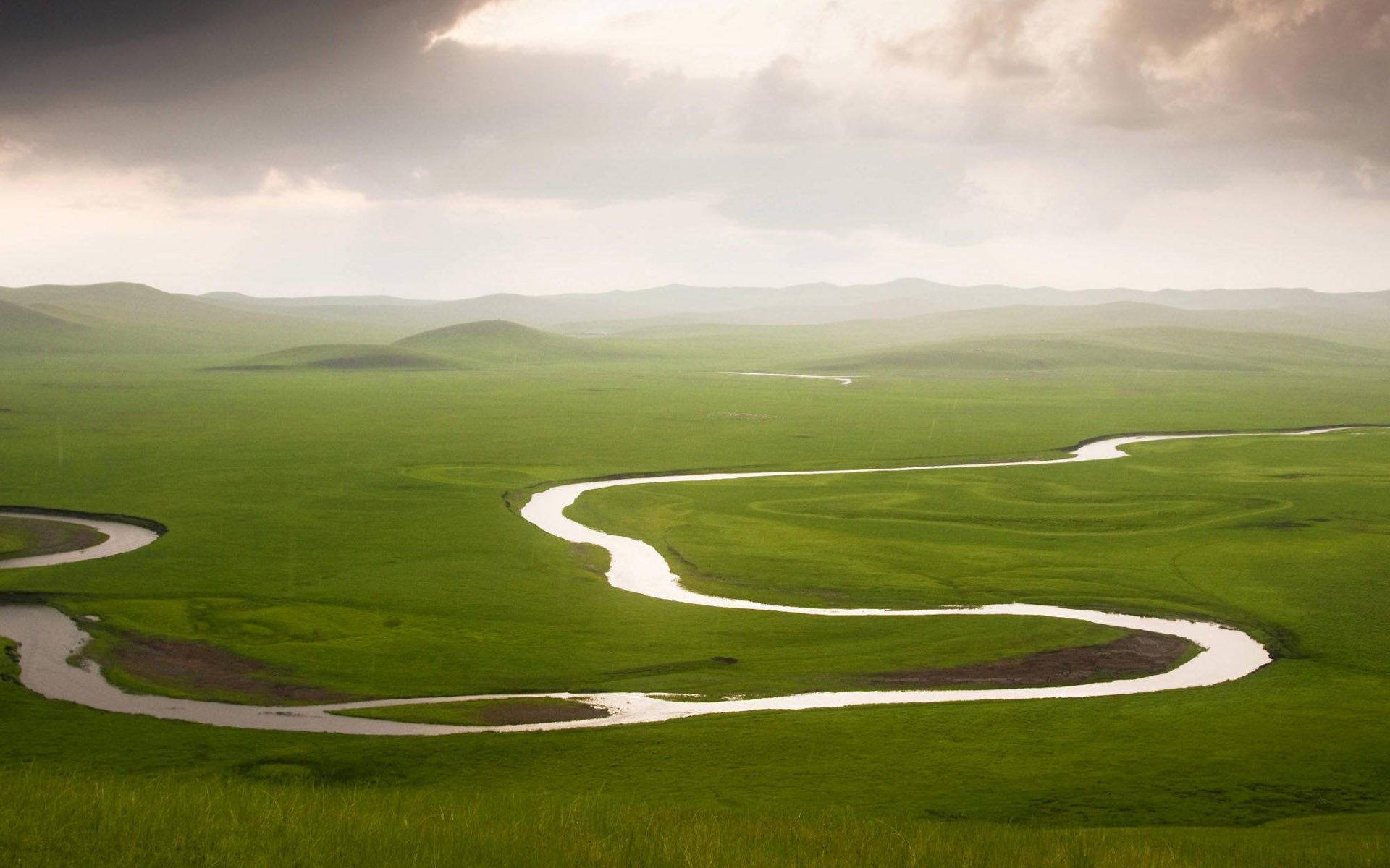 Winding river through green rolling hills under cloudy sky HD Wallpaper