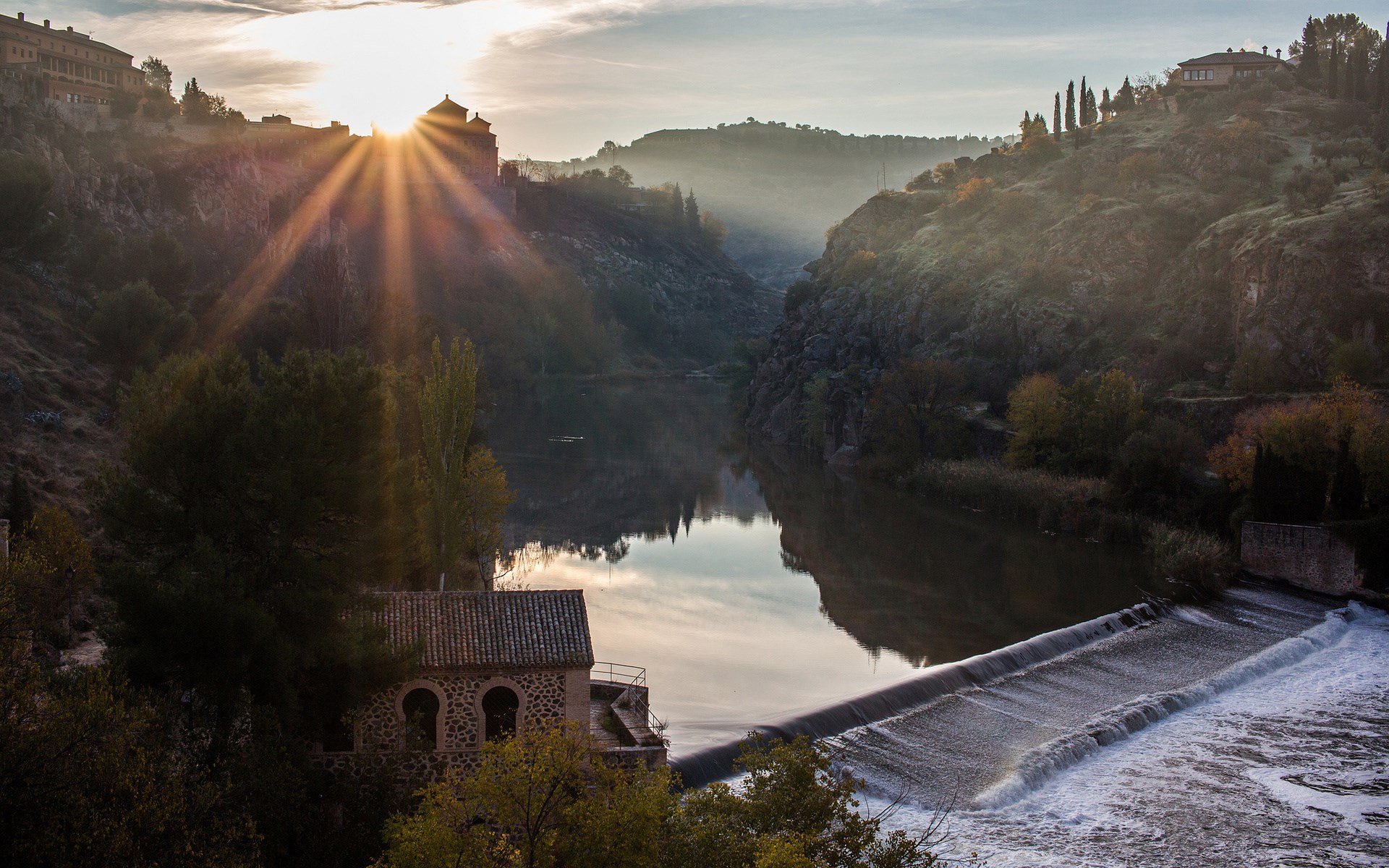 River flowing past a dam with sunbeams over a town HD Wallpaper