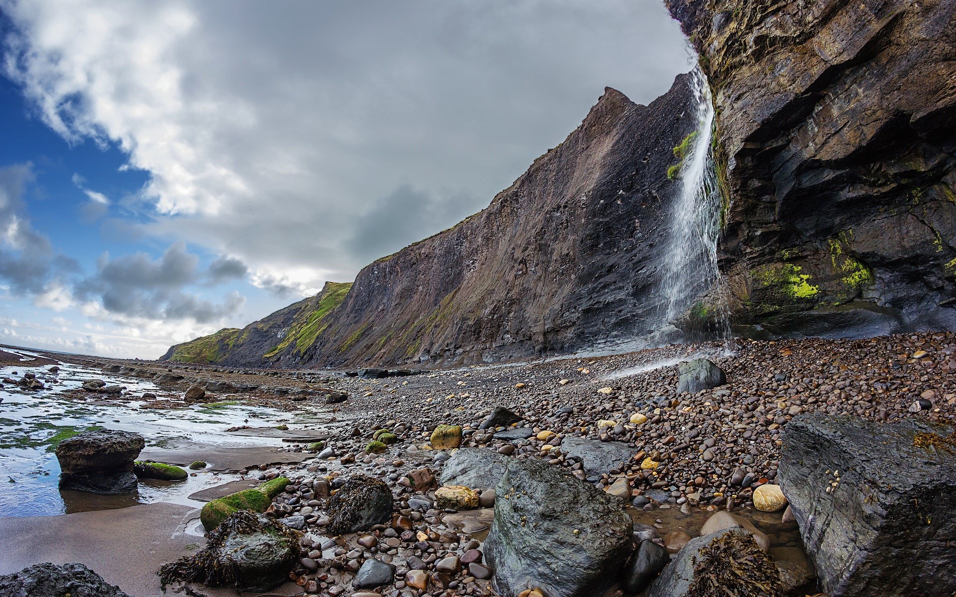 Waterfall cascading down a rocky cliff face onto a pebble beach HD Wallpaper