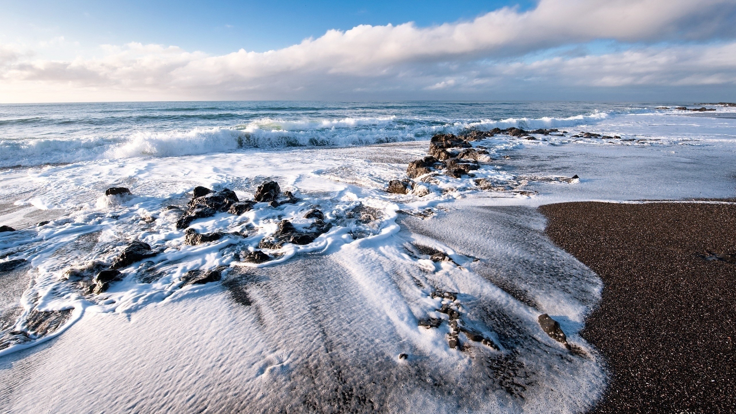 Waves crashing on a rocky, dark sand beach HD Wallpaper