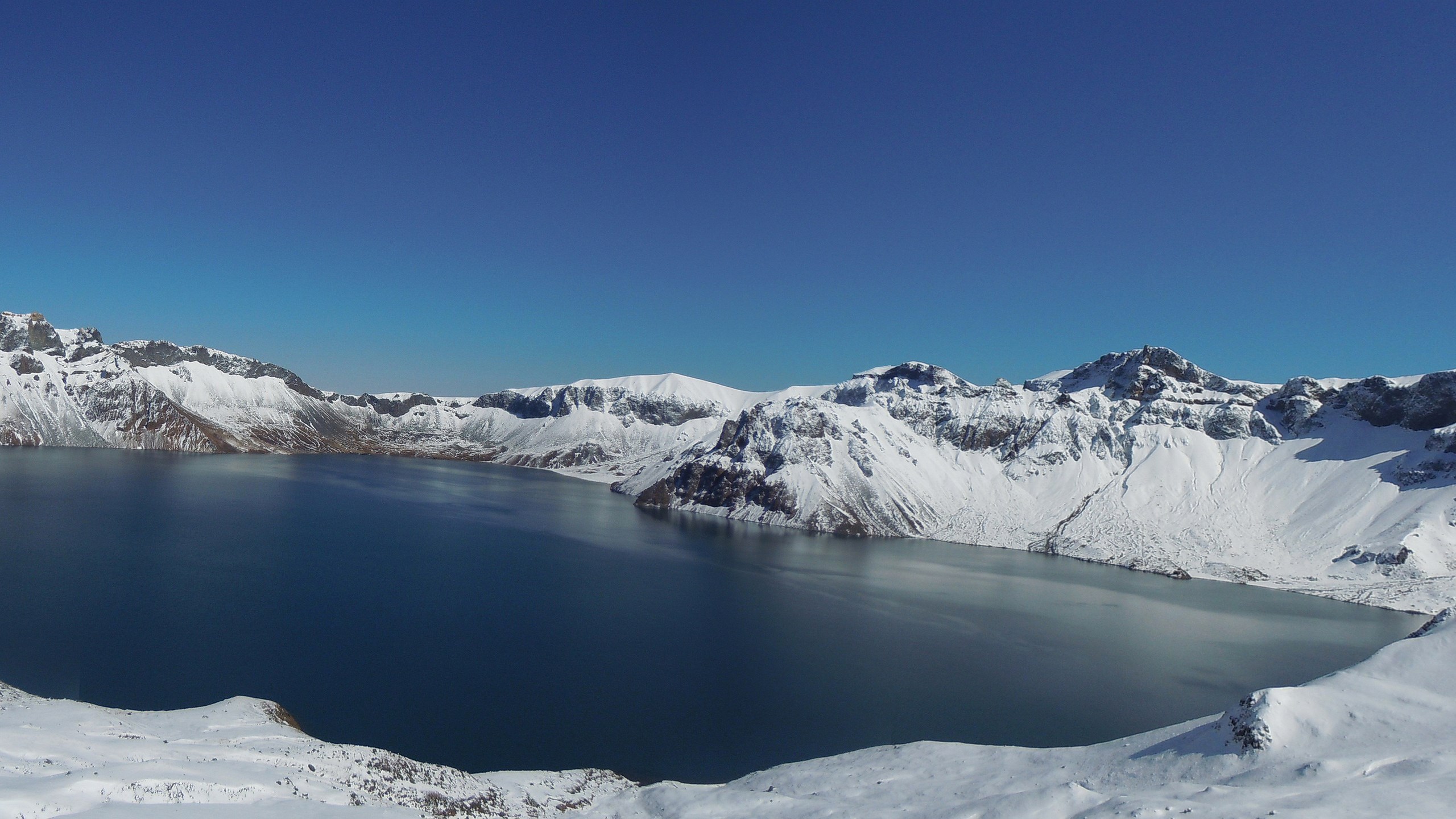 Snow-covered mountains surrounding a deep blue lake under a clear sky HD Wallpaper