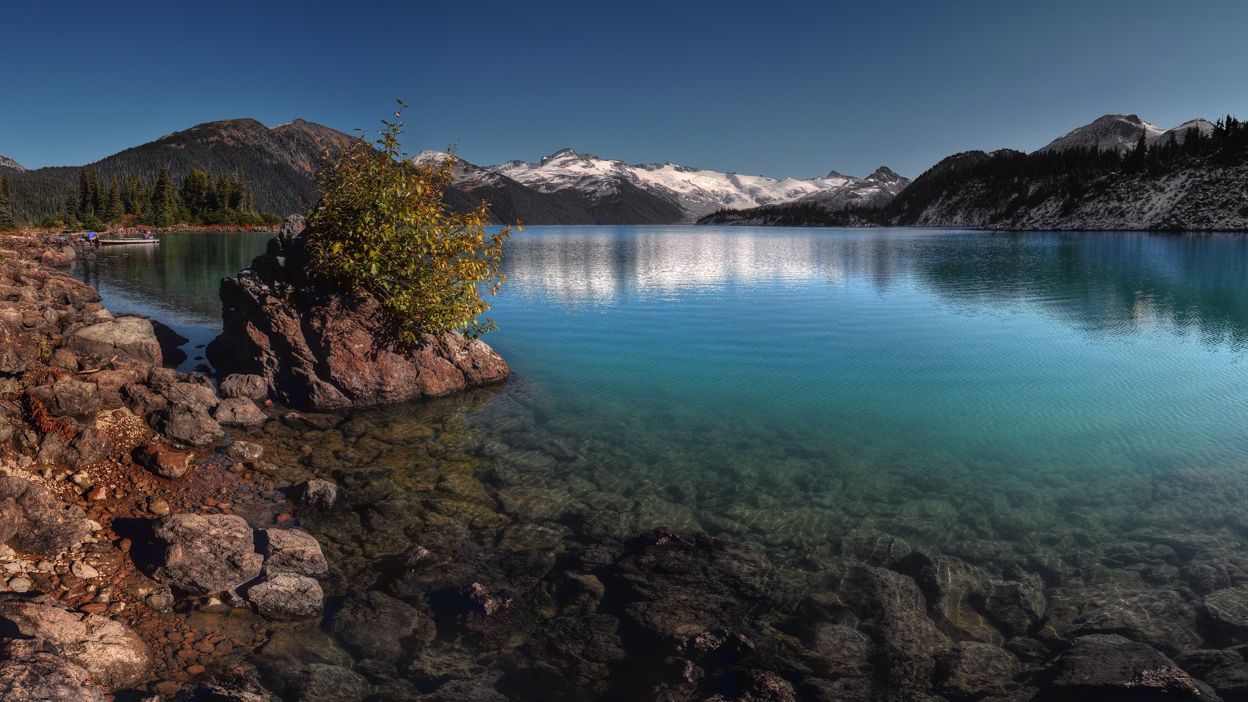 Turquoise lake with snow-capped mountains and rocky shore HD Wallpaper