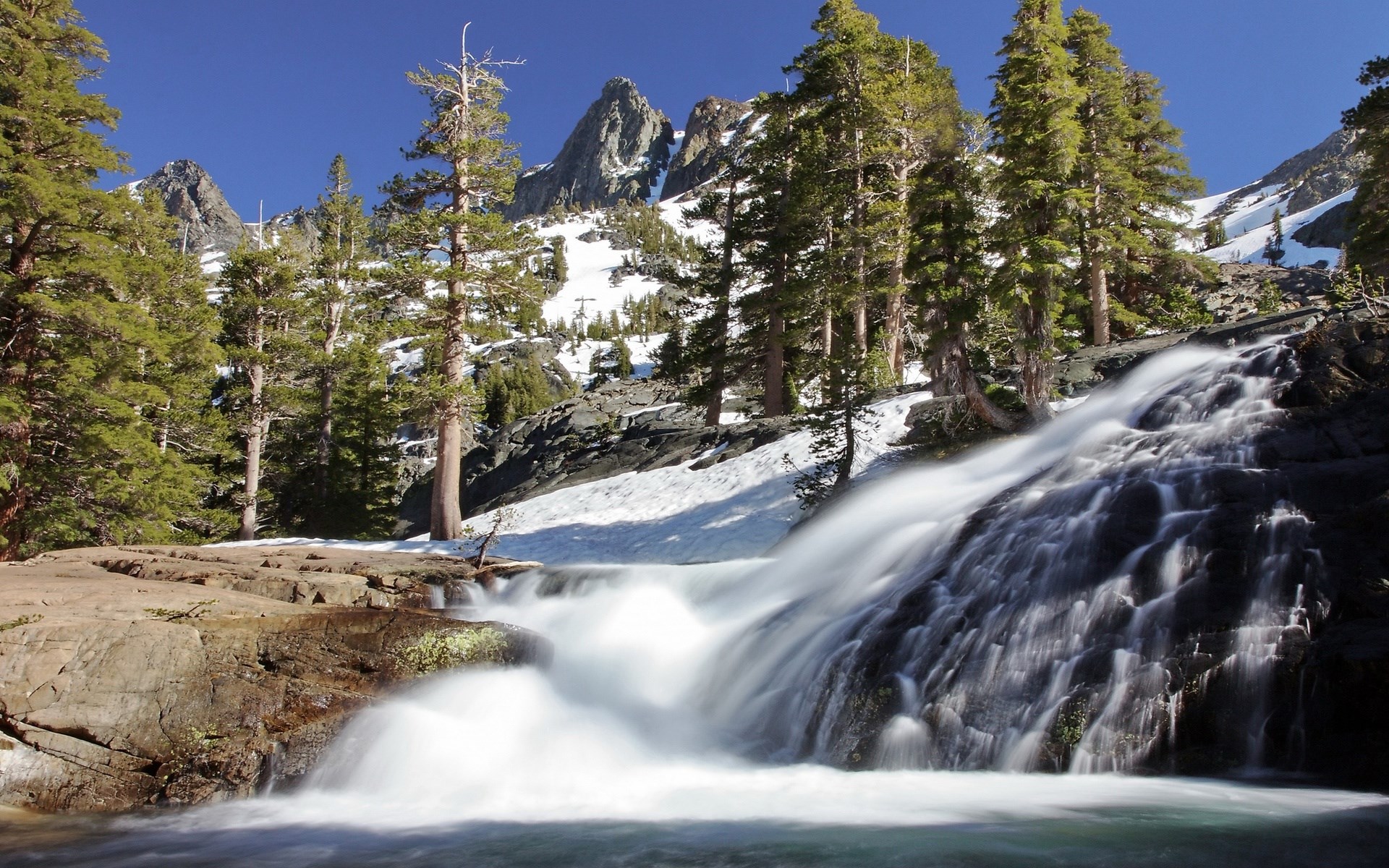 Waterfall flowing over rocks with pine trees and snowy mountains HD Wallpaper
