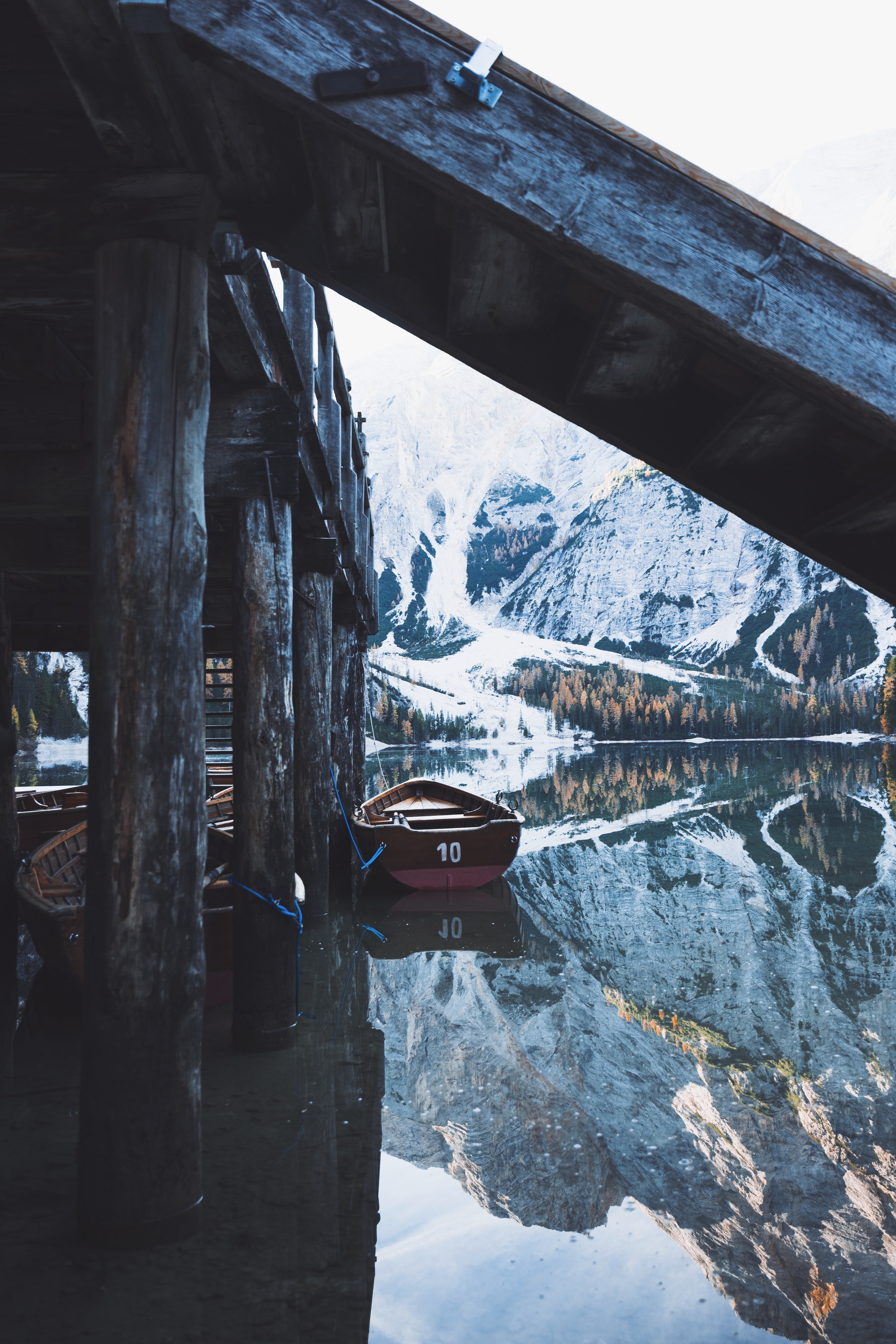 Boats docked by a wooden structure with snowy mountains reflected in the water HD Wallpaper