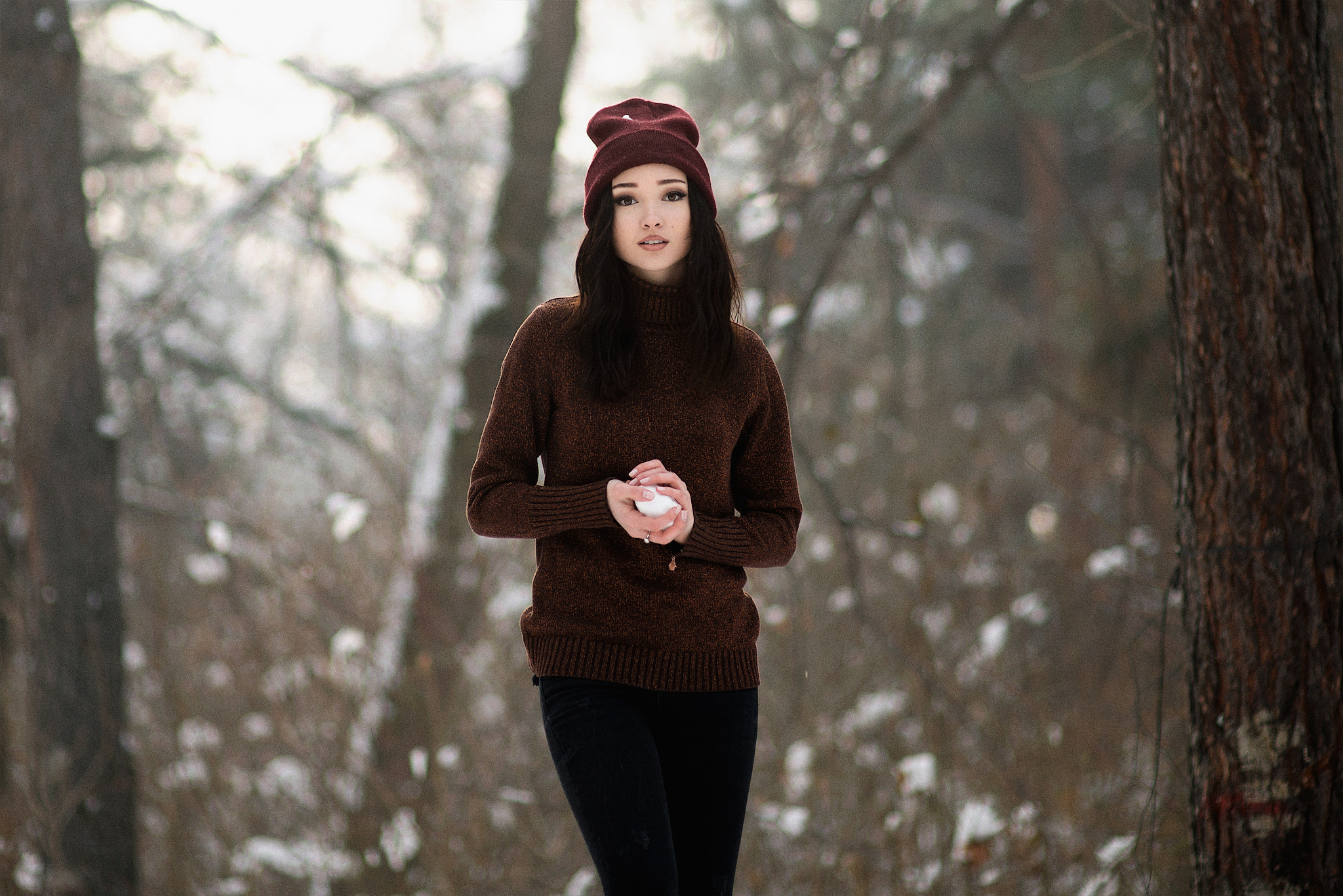 Woman in a maroon beanie and sweater holding a snowball in a snowy forest HD Wallpaper