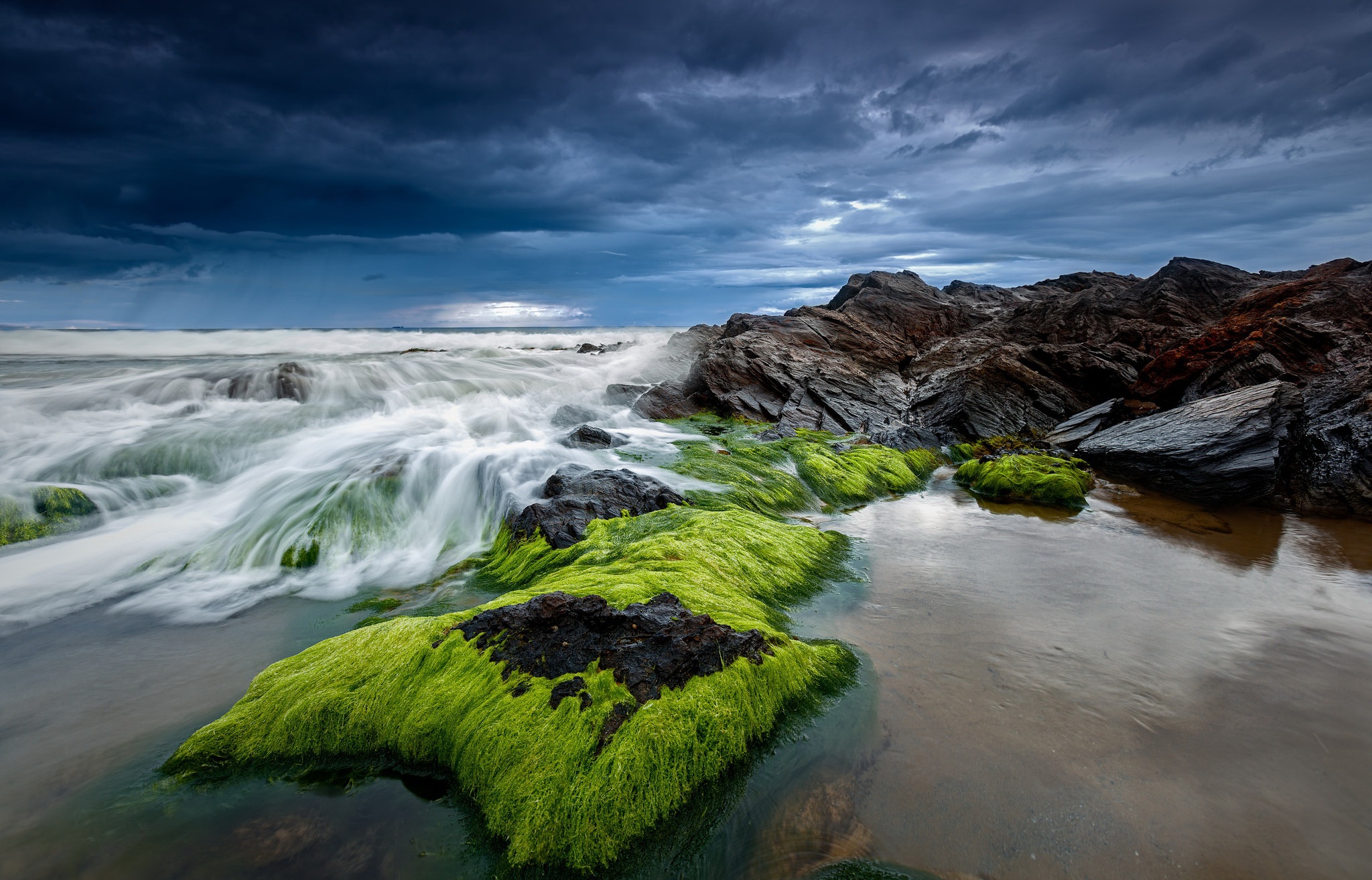 Waves crashing on moss-covered rocks under a stormy sky HD Wallpaper