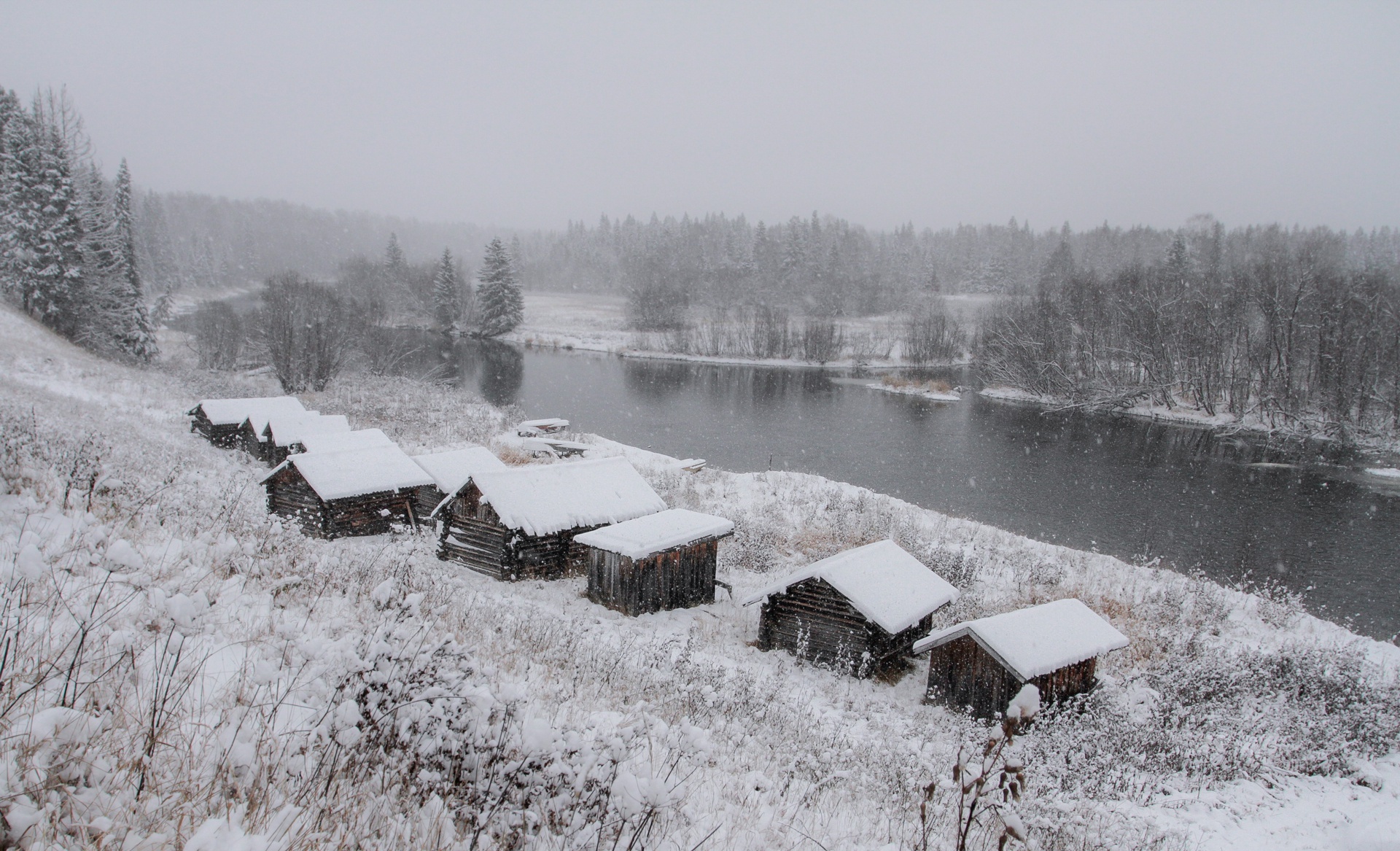 Wooden cabins by a river during a snowfall HD Wallpaper