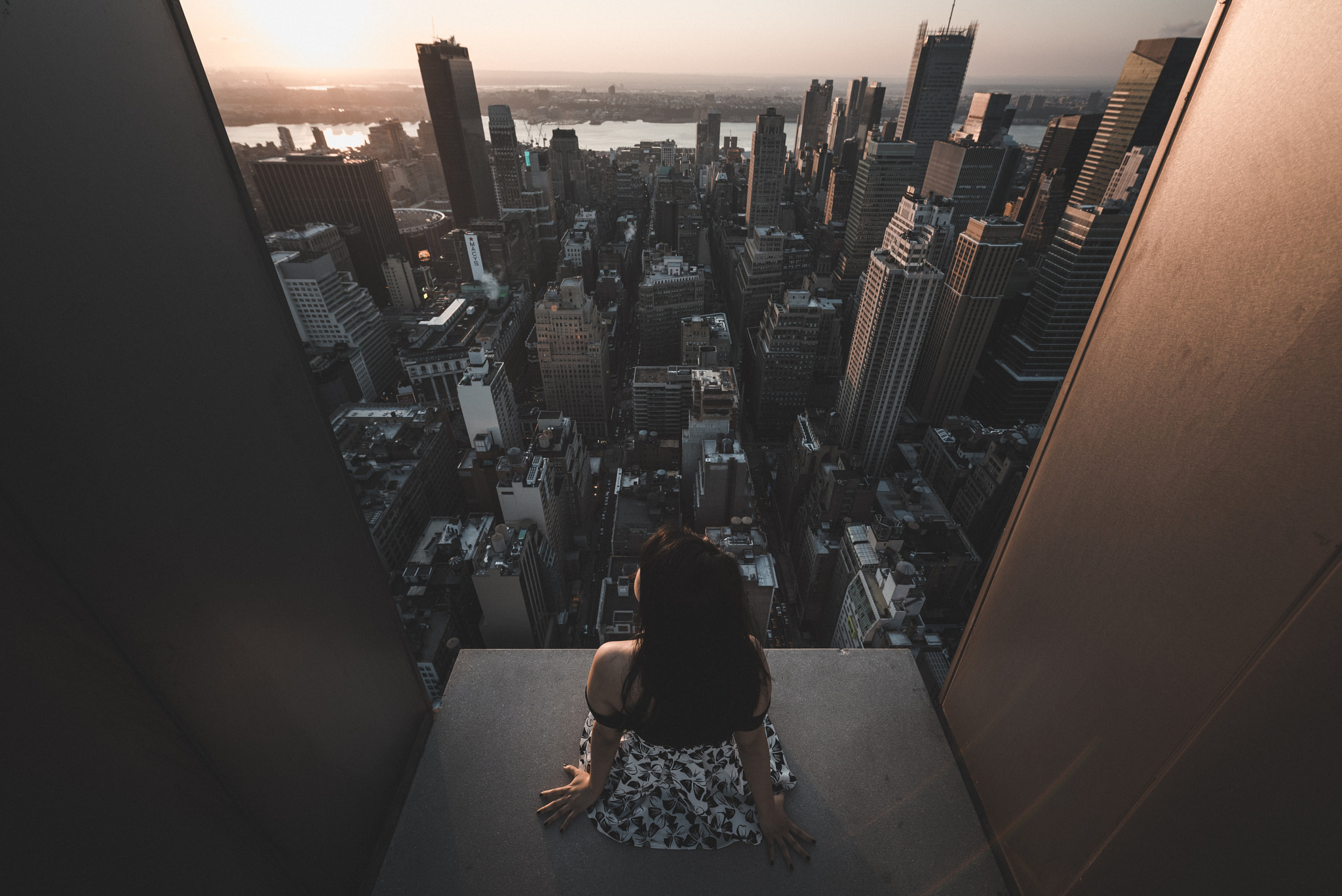 Woman sitting on edge of skyscraper overlooking cityscape at sunset HD Wallpaper