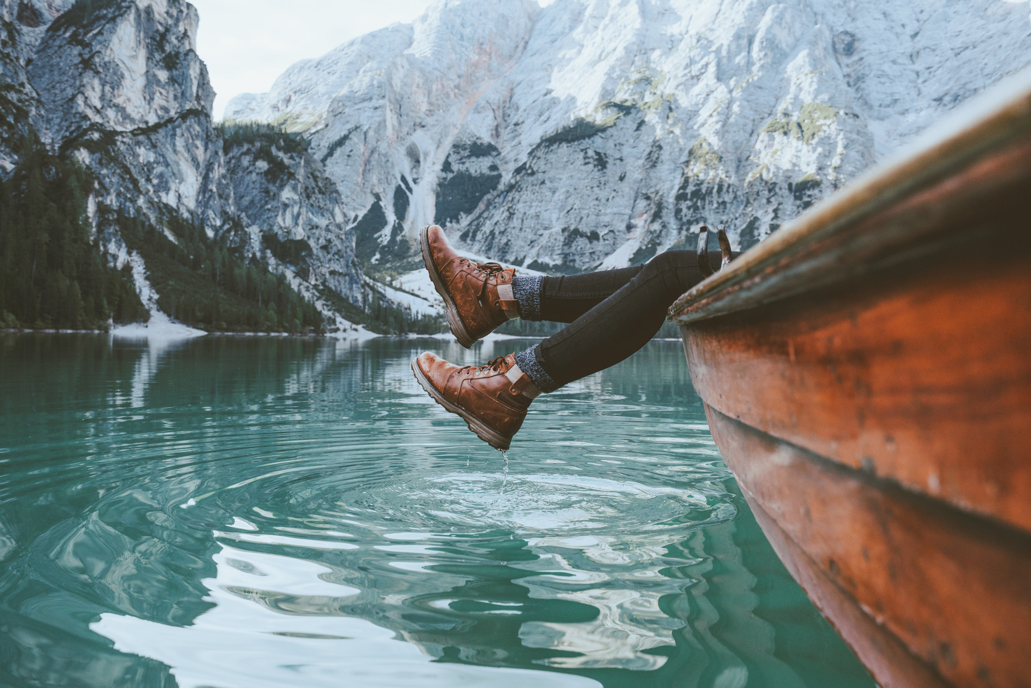 Person's legs dangling from a boat on a lake with mountains HD Wallpaper
