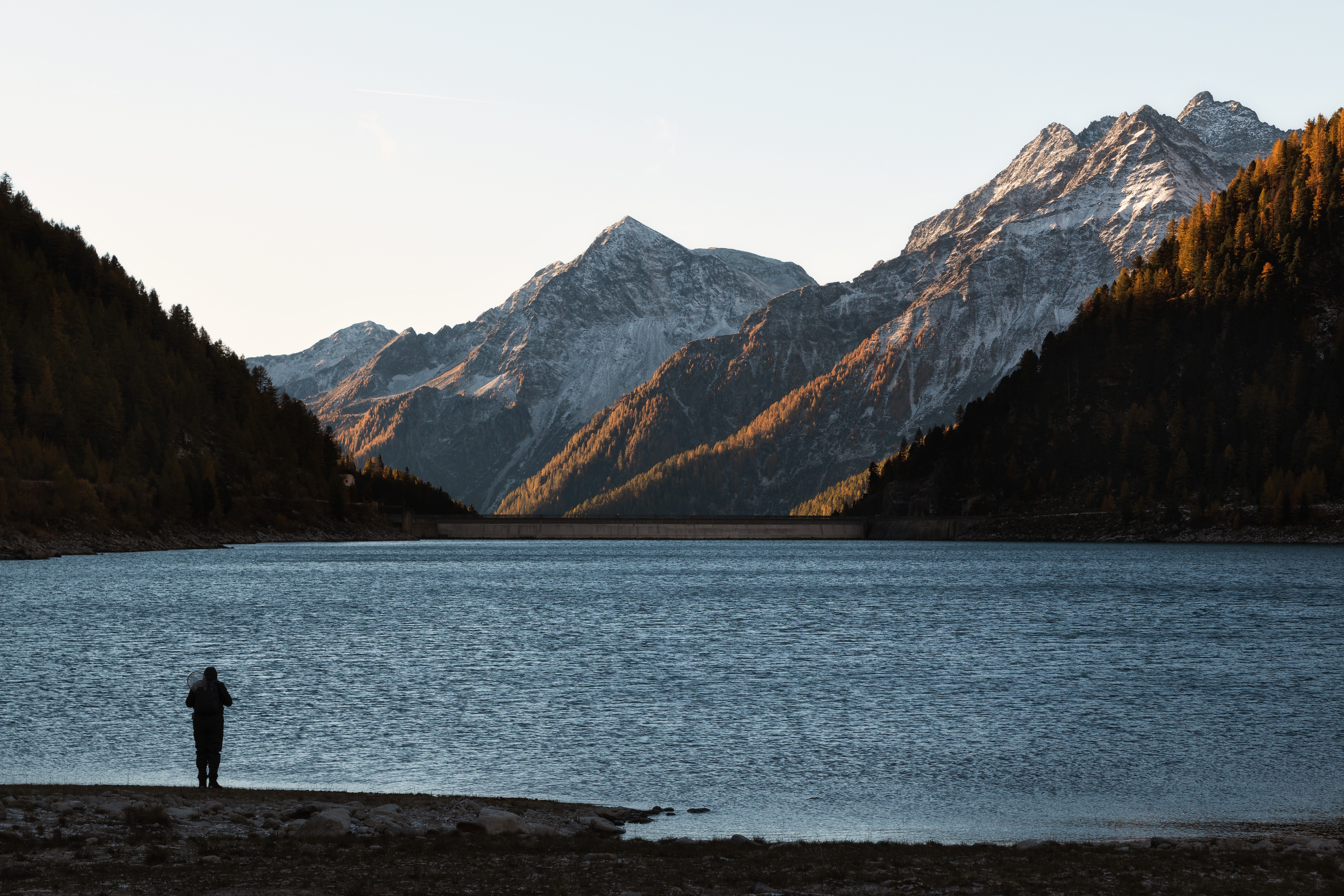 Person standing by a lake with mountains and a dam in the background HD Wallpaper
