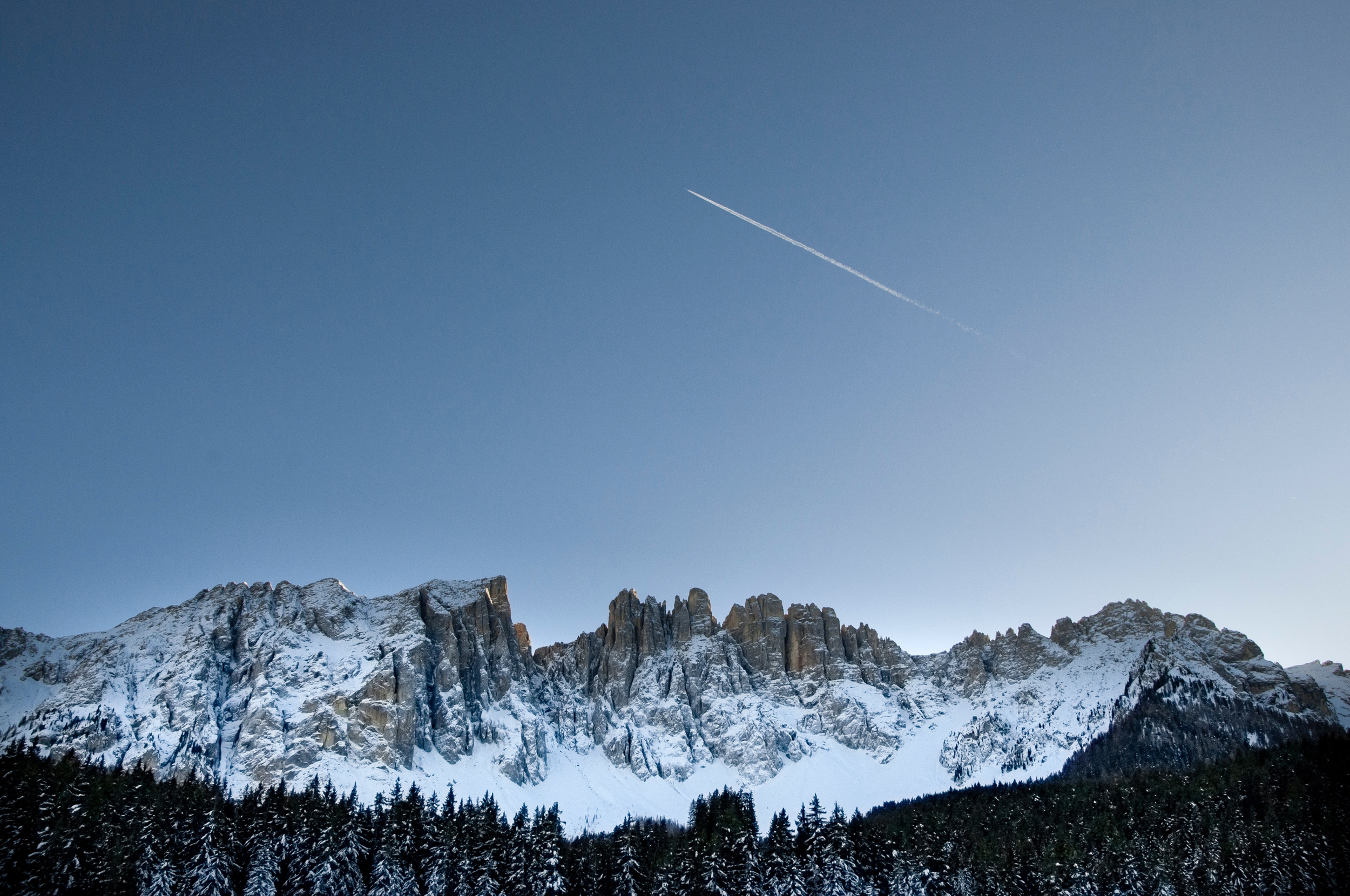 Snowy mountains with pine trees and a contrail in the sky HD Wallpaper