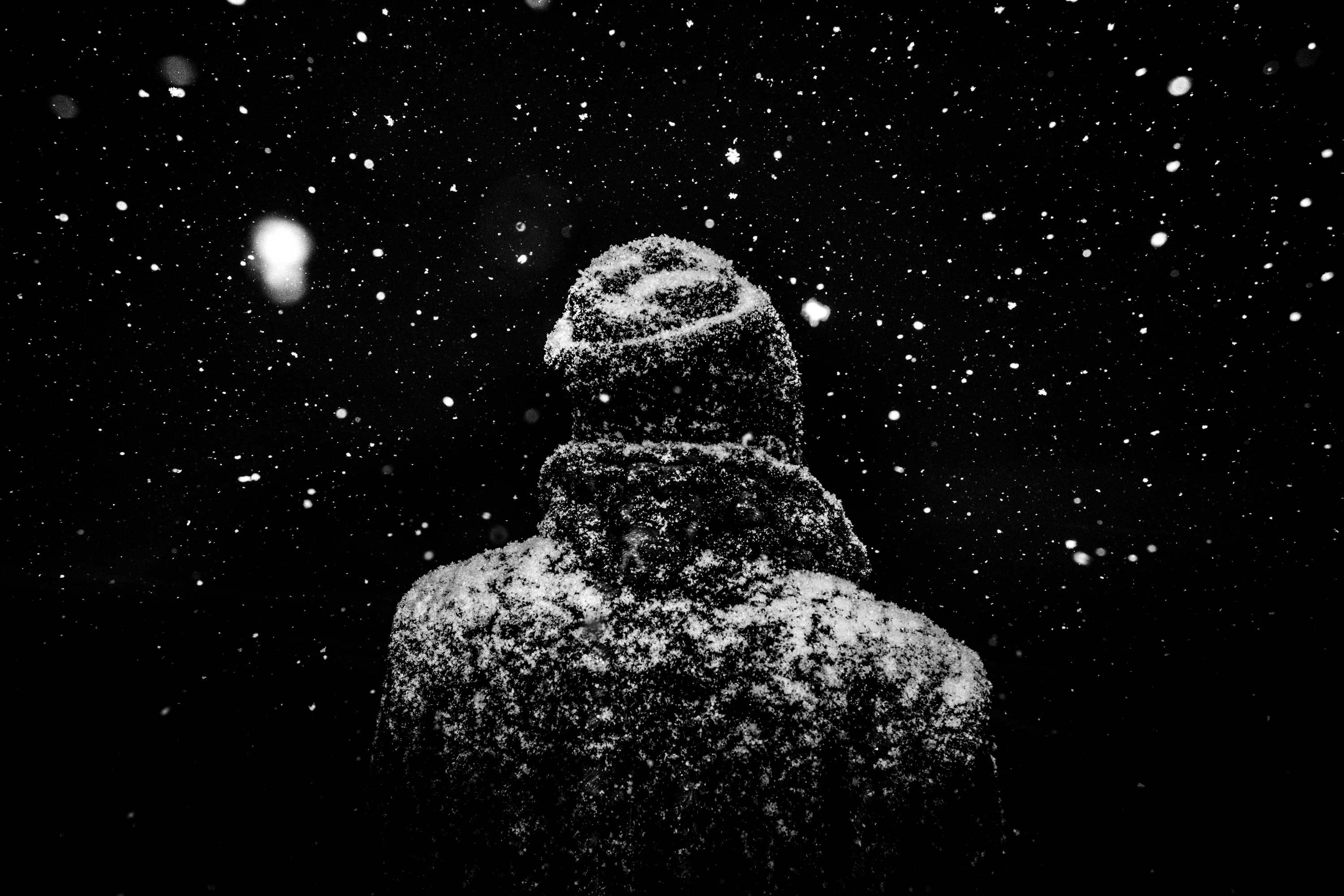 Man in a snow-covered coat and beanie during snowfall HD Wallpaper