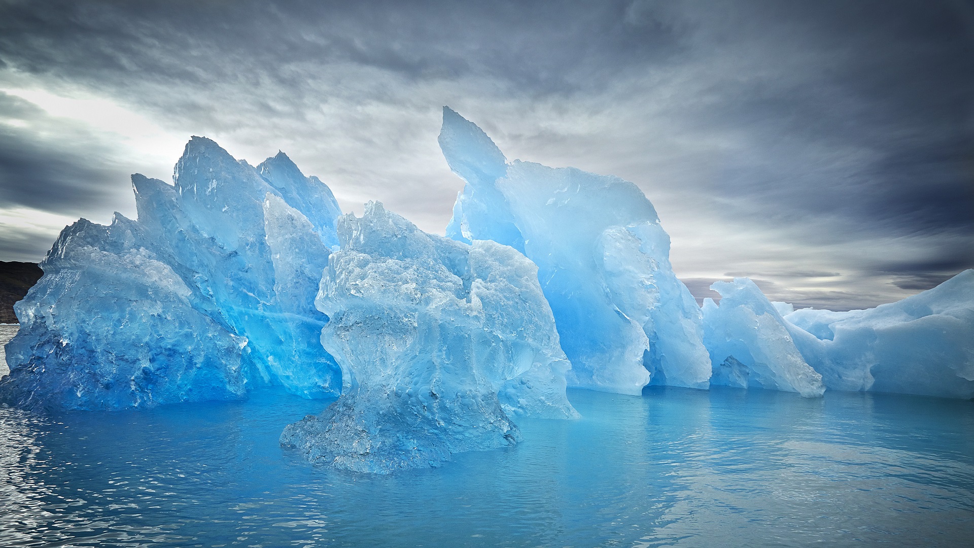Blue iceberg floating in bright blue water under cloudy sky HD Wallpaper