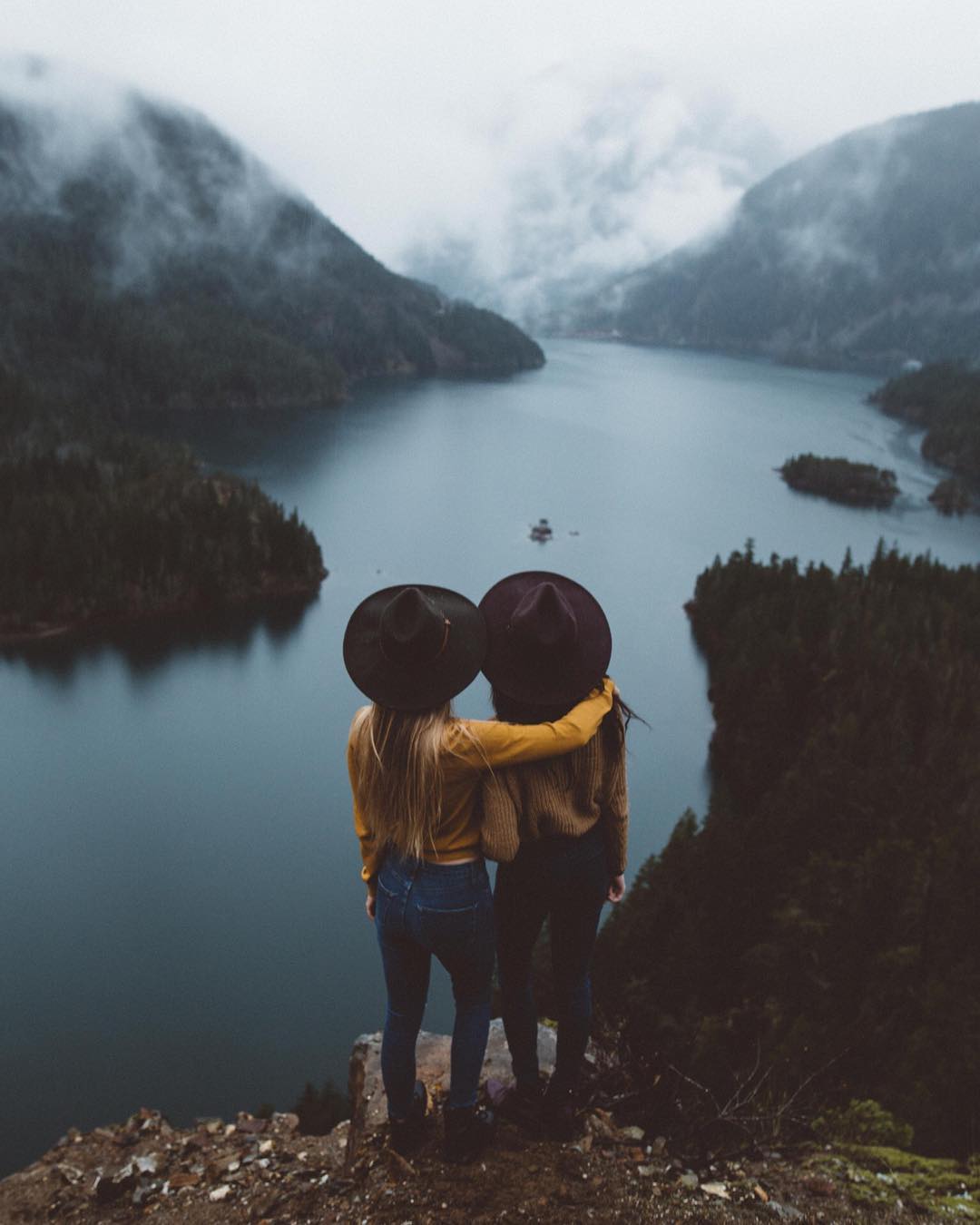 Two women in hats hugging on a cliff overlooking a lake HD Wallpaper