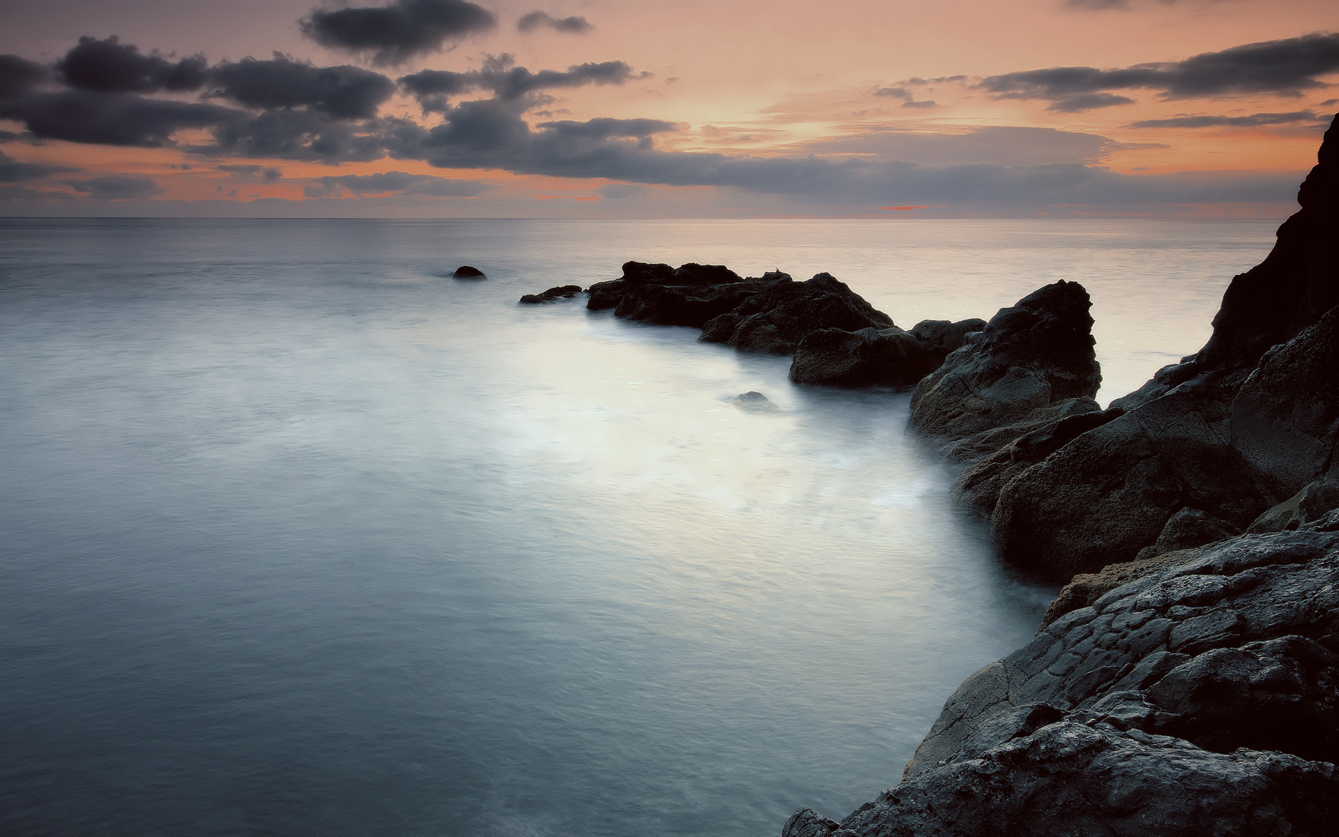 Rocky coastline at sunset with calm sea HD Wallpaper