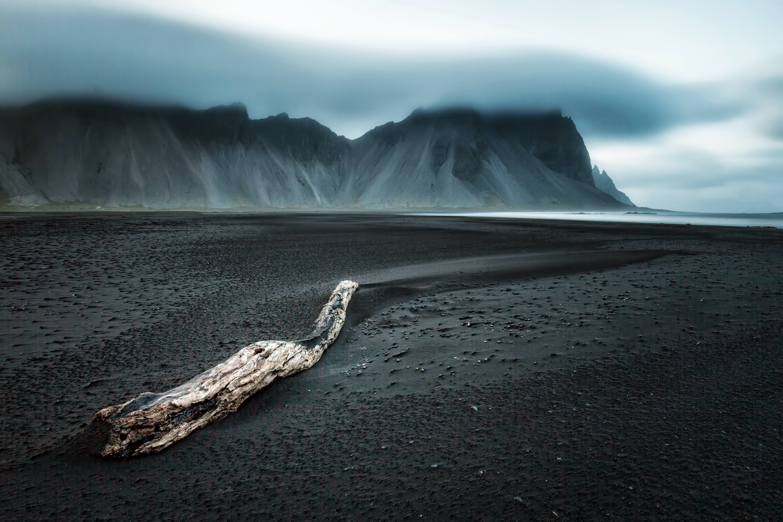Driftwood on a black sand beach with mountains HD Wallpaper