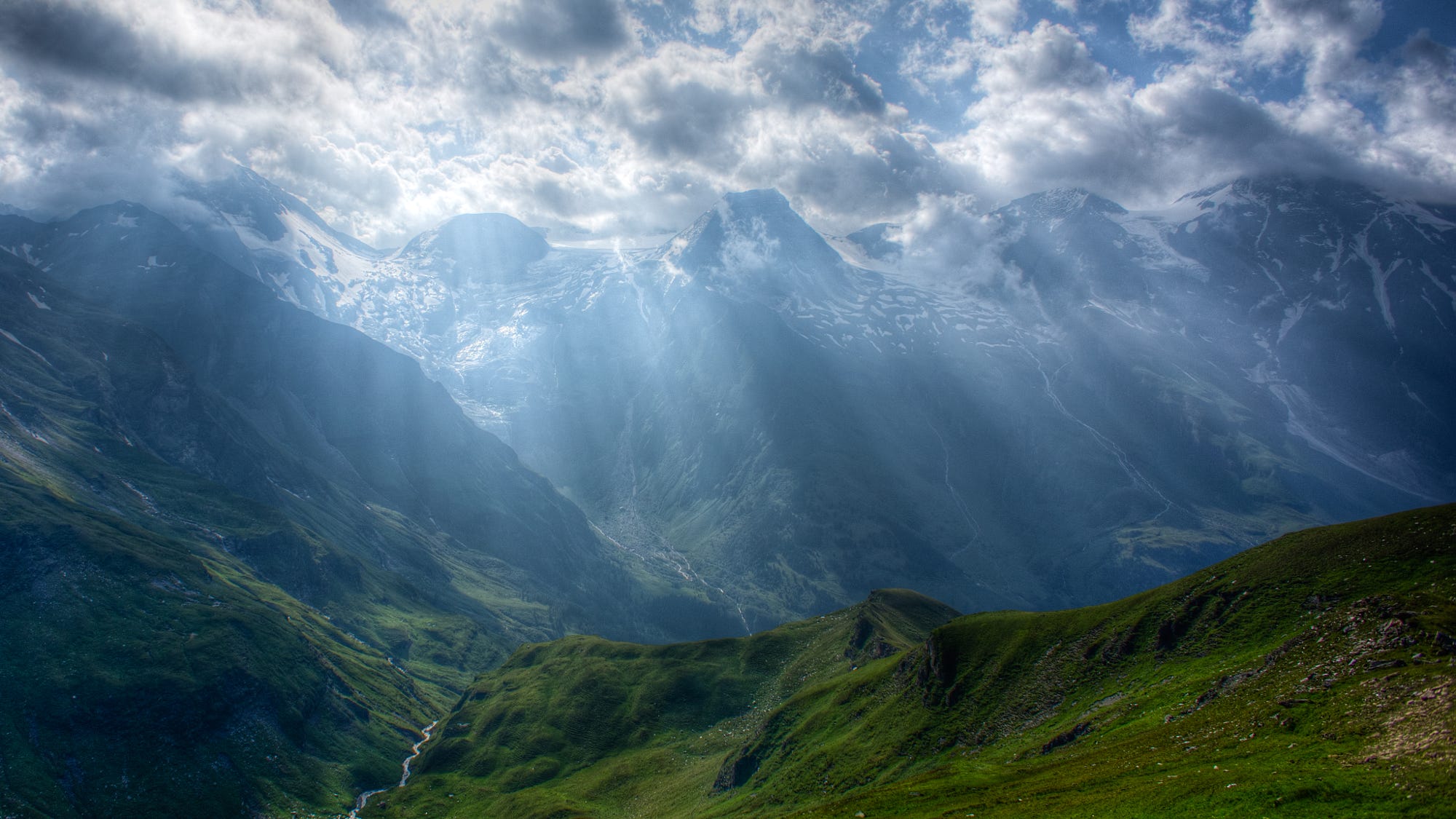 Sun rays over snow-capped mountains and green valley HD Wallpaper