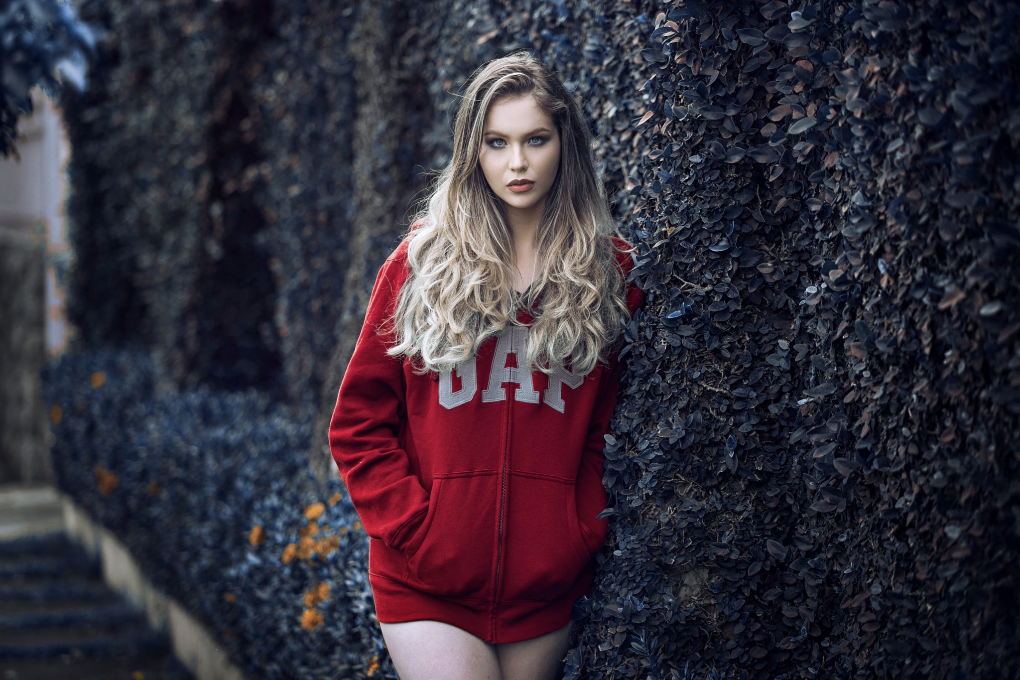 Woman in red GAP hoodie leaning against a bush HD Wallpaper