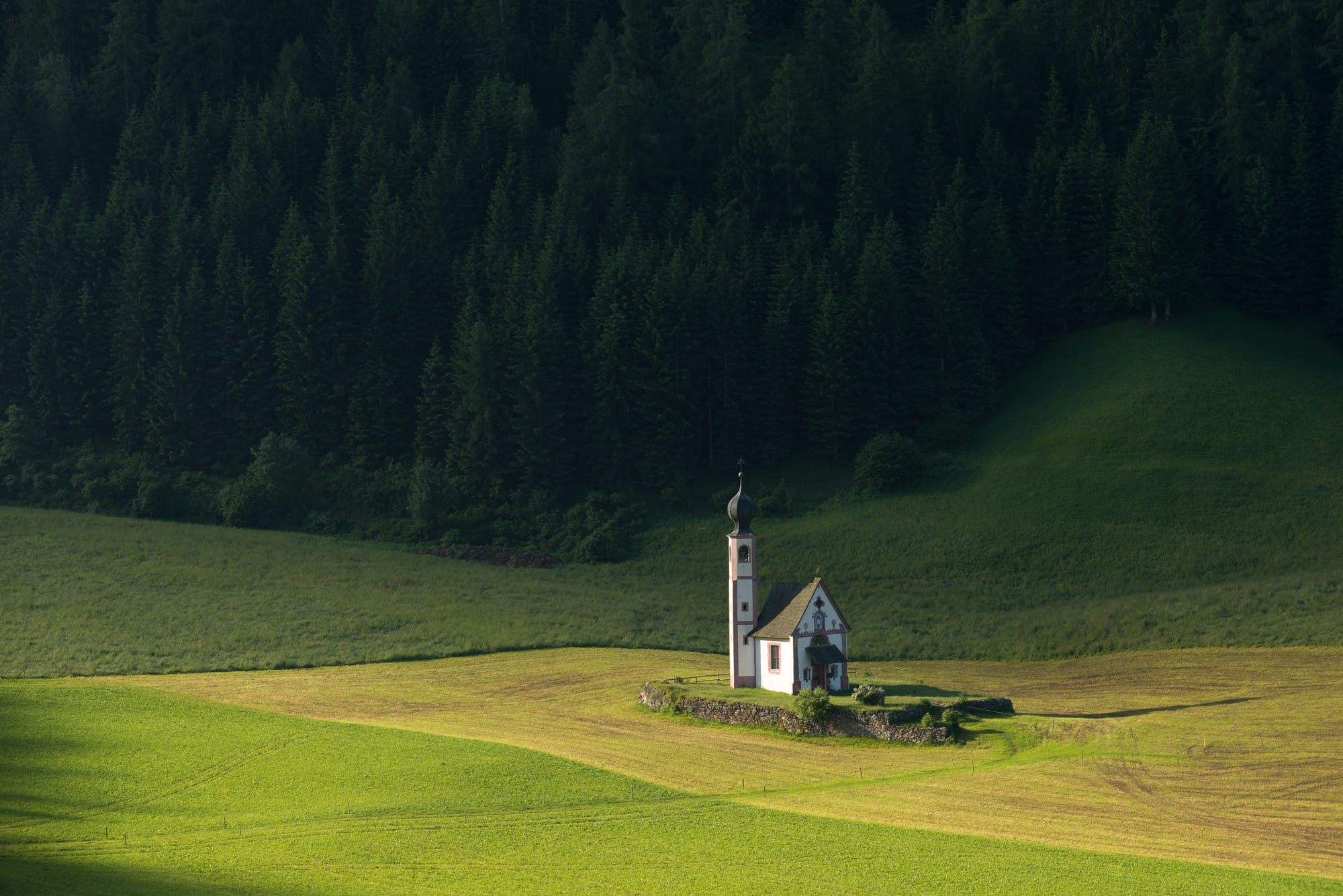 A small church stands alone in a green field with a forest behind it. HD Wallpaper