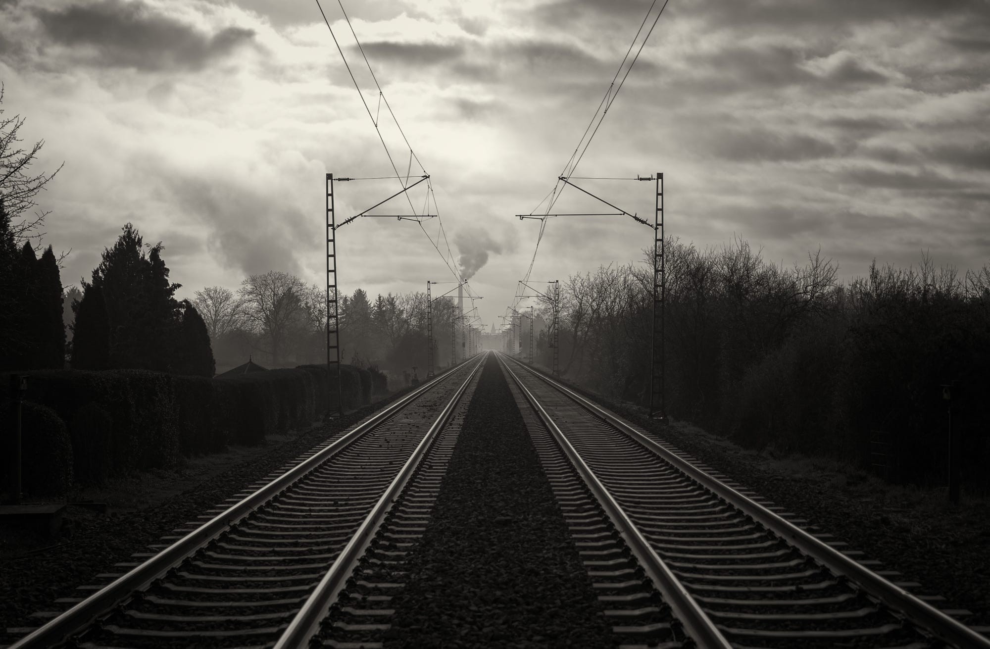 Railway tracks leading to a smoking industrial chimney under cloudy sky HD Wallpaper