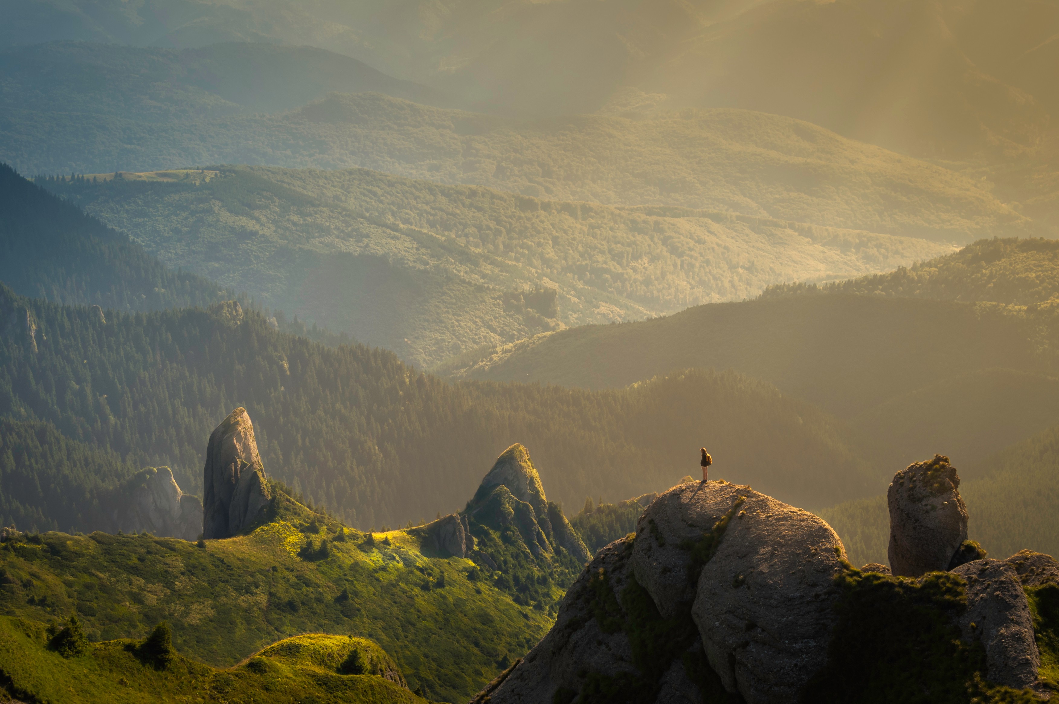Hiker on a rocky peak overlooking a forested mountain range at sunrise HD Wallpaper