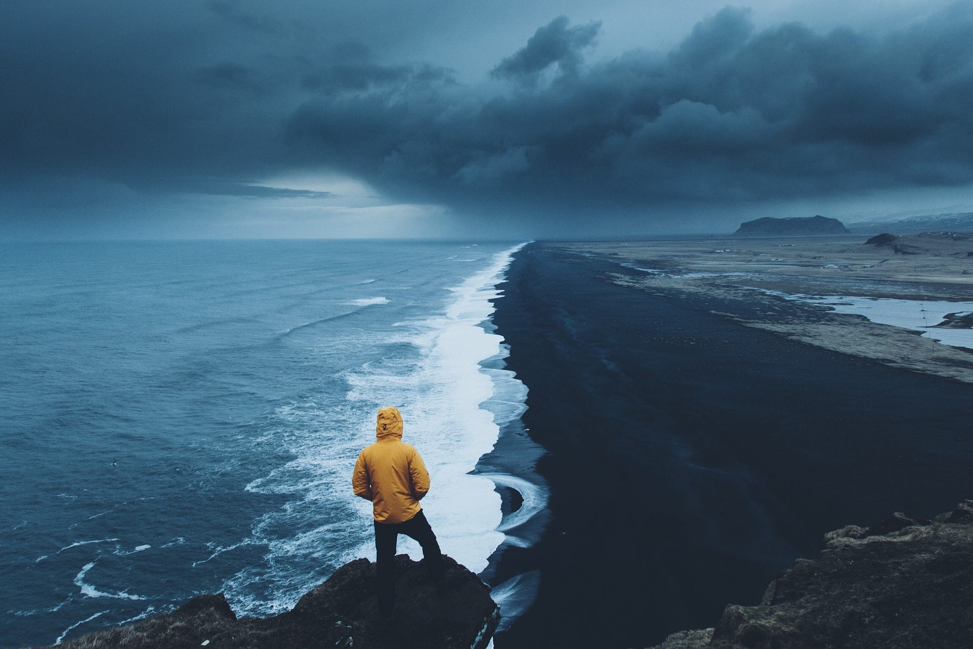 Person in yellow jacket on cliff overlooking black sand beach and ocean HD Wallpaper